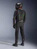 Rider from behind wearing Alpinestars GP Force V2 Leather Suit, 1-Piece, Black with Green accents, aerodynamic speed hump, paired with a black carbon-fiber helmet, black racing boots, and gloves, high-performance motorcycle racing gear