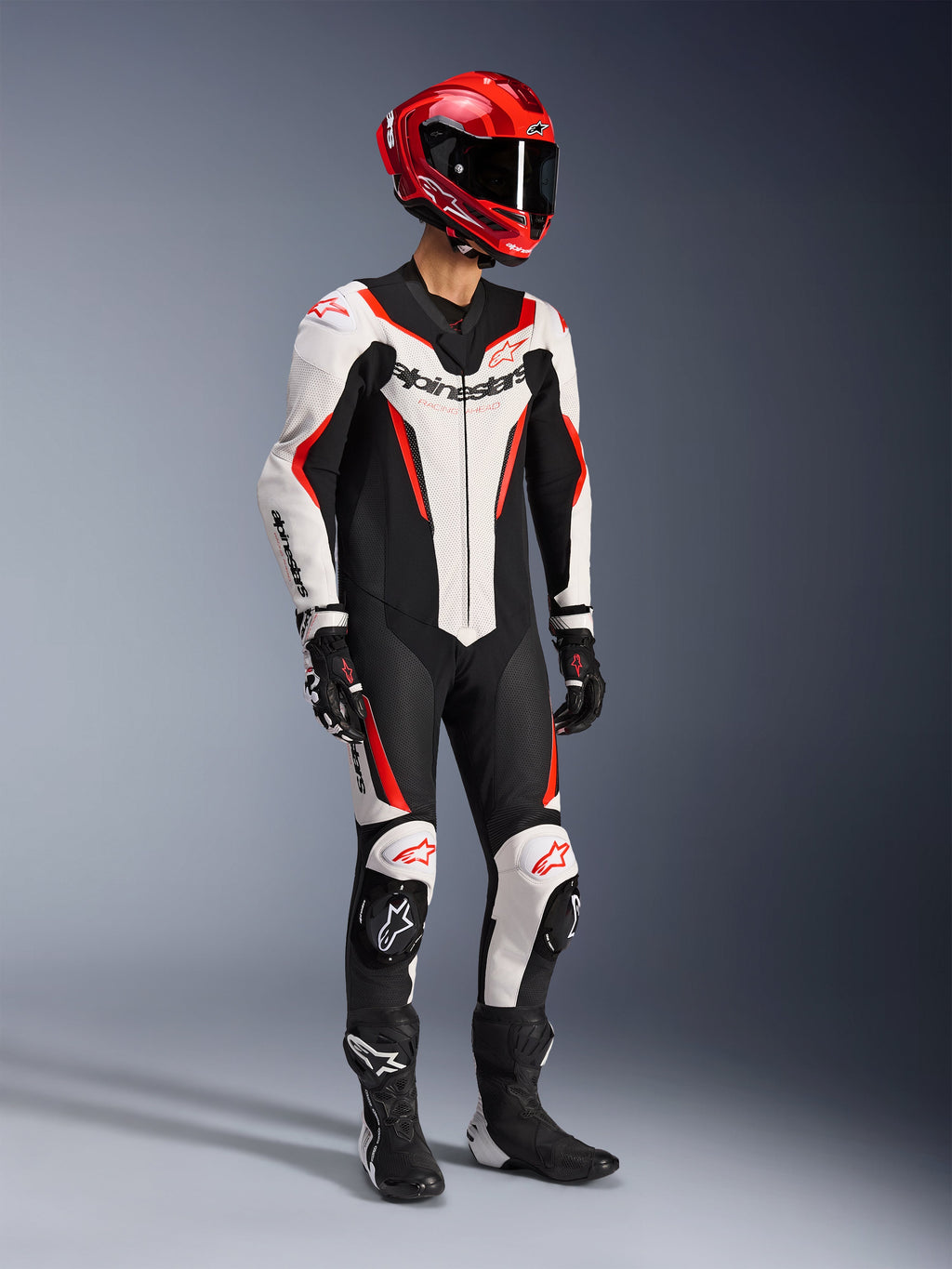 Combinaison en cuir Alpinestars GP Force V2 1 pièce en blanc, noir et rouge fluo, photo de studio en plein pied d'un pilote portant un équipement de course complet incluant un casque rouge, des gants noirs et des bottes noires, dotée d'une construction aérodynamique en cuir bovin avec marquage distinctif et sliders de genoux intégrés.