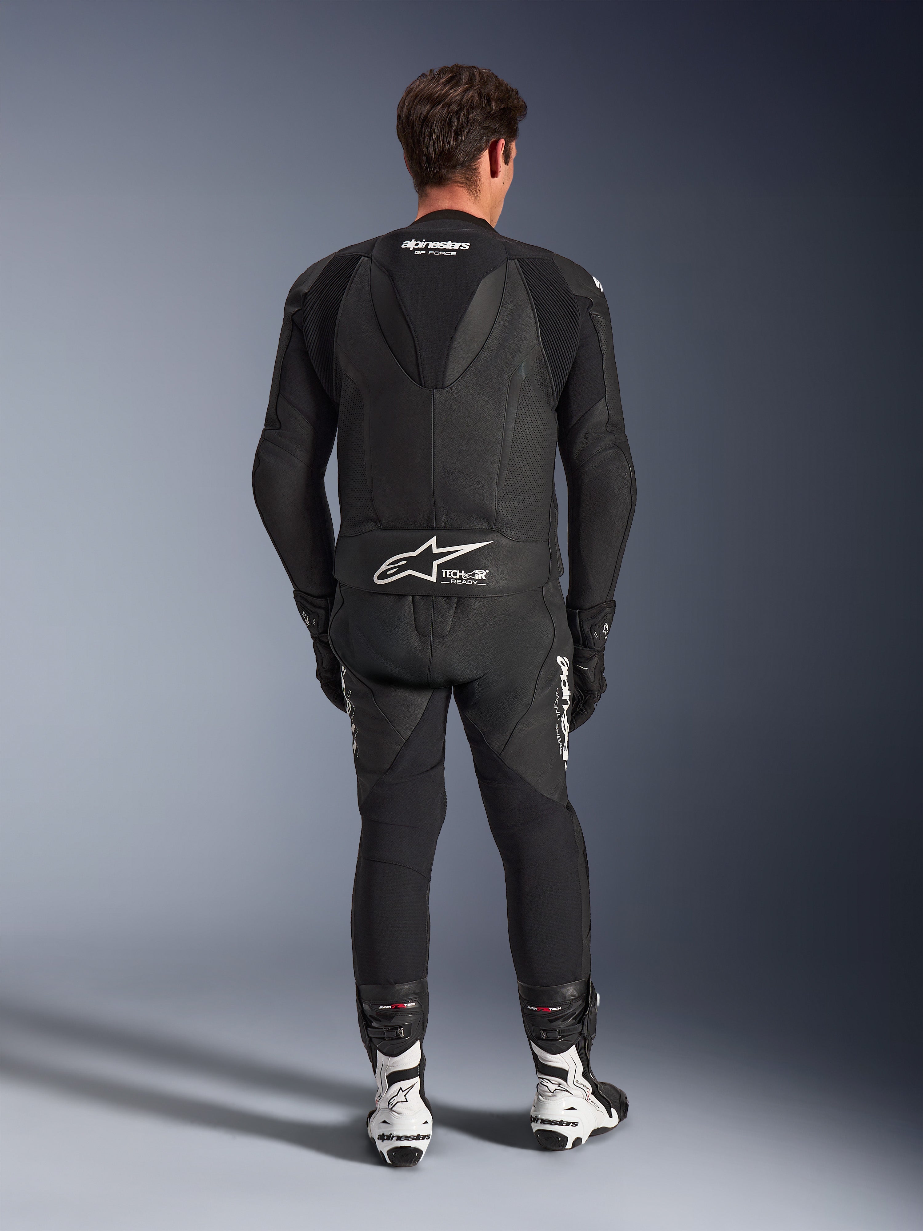 Combinaison en cuir Alpinestars GP Force V2 2 pièces, noire, vue arrière montrant la bosse aérodynamique et le marquage Tech-Air Ready à la taille, panneaux en cuir perforé pour la ventilation, assortie à des bottes de course blanches et noires, équipement de course de moto professionnel