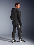 Modèle masculin portant la combinaison en cuir Alpinestars GP Force V2 2 pièces, noir avec logos blancs, vue de trois-quarts arrière montrant la bosse aérodynamique et le marquage Tech-Air Ready, avec bottes et gants de course noirs et blancs, équipement de moto professionnel pour sport et piste