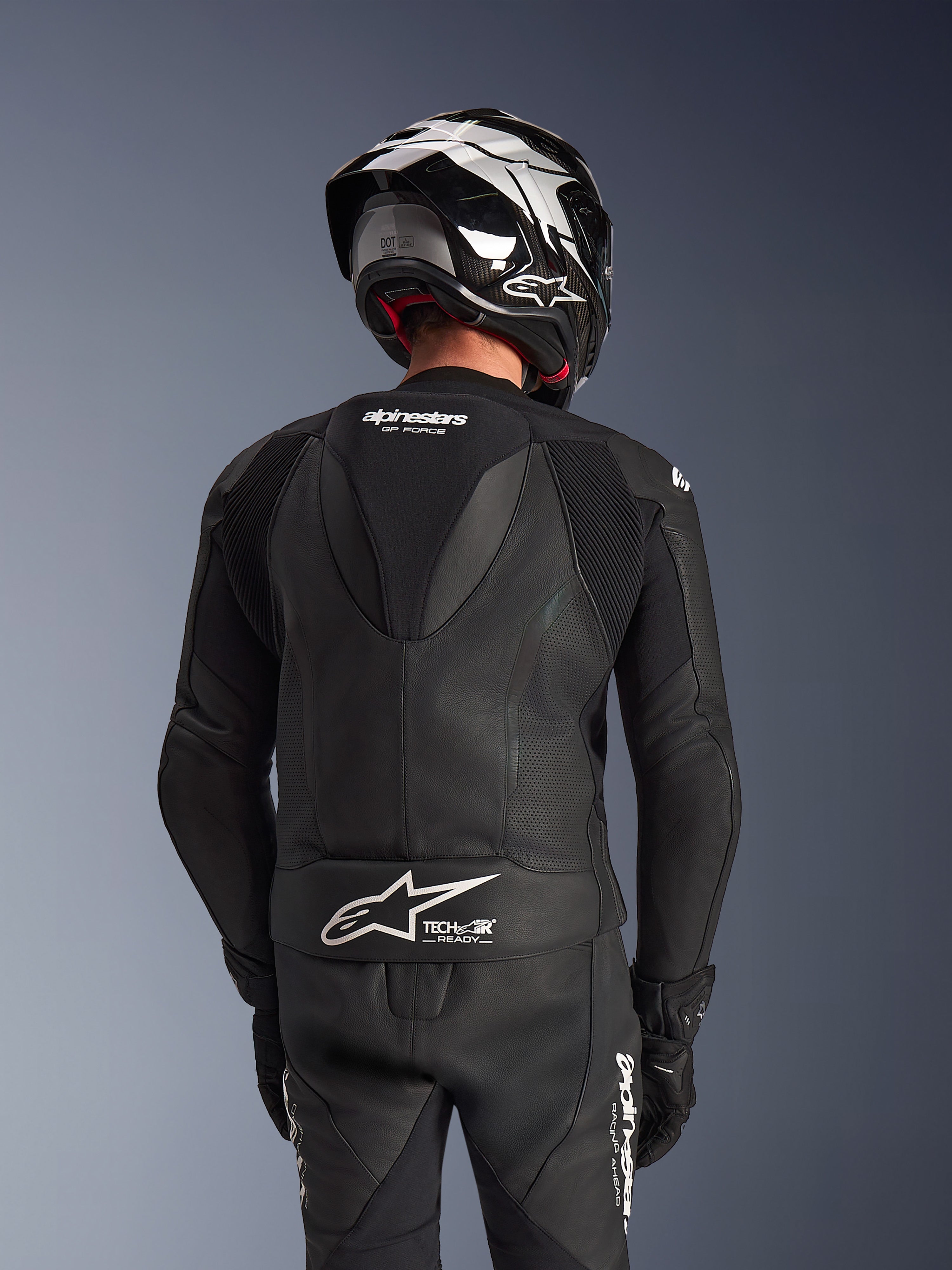 Combinaison en cuir Alpinestars GP Force V2 2 pièces, noire, vue arrière du pilote montrant la bosse aérodynamique, les panneaux en cuir perforé et les zones extensibles en accordéon sur les épaules, avec marquage Tech-Air Ready dans le bas du dos, portée avec un casque de course et des gants assortis.