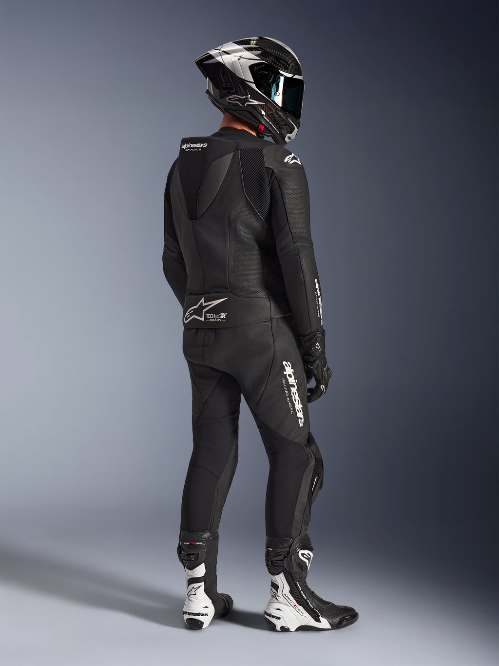 Combinaison en cuir 2 pièces Alpinestars GP Force V2, noire, vue arrière d'un pilote portant un équipement de course complet, avec bosse aérodynamique, logos blancs et cuir perforé, associée à un casque en fibre de carbone et des bottes de course noires et blanches