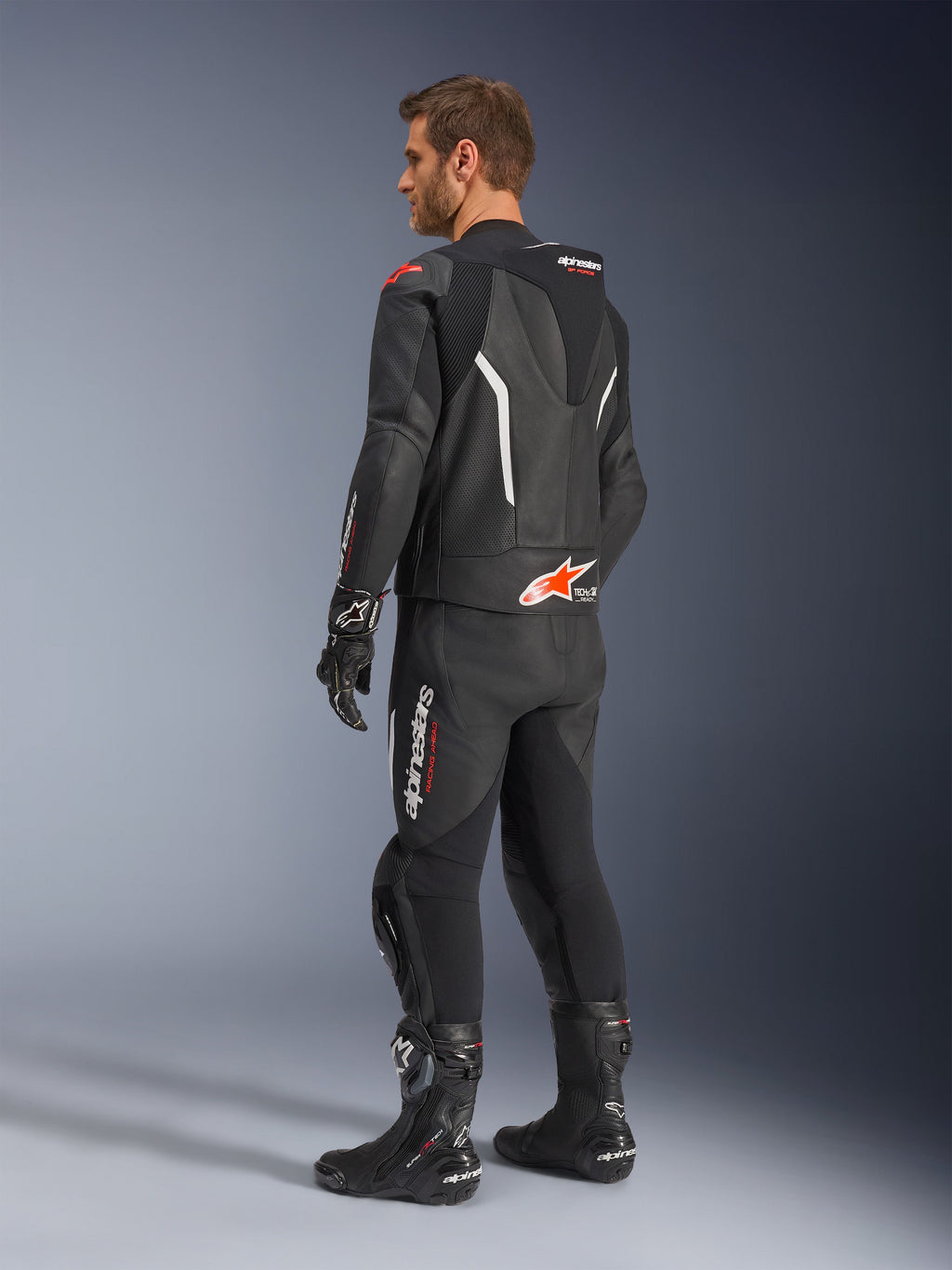 Alpinestars GP Force V2 2-teiliger Lederanzug, Schwarz mit weißen und Red Fluo Akzenten, Dreiviertel-Rückansicht am Modell, mit aerodynamischem Höcker, perforierten Ledereinsätzen und markantem Alpinestars-Branding, getragen mit schwarzen Rennstiefeln und Handschuhen.