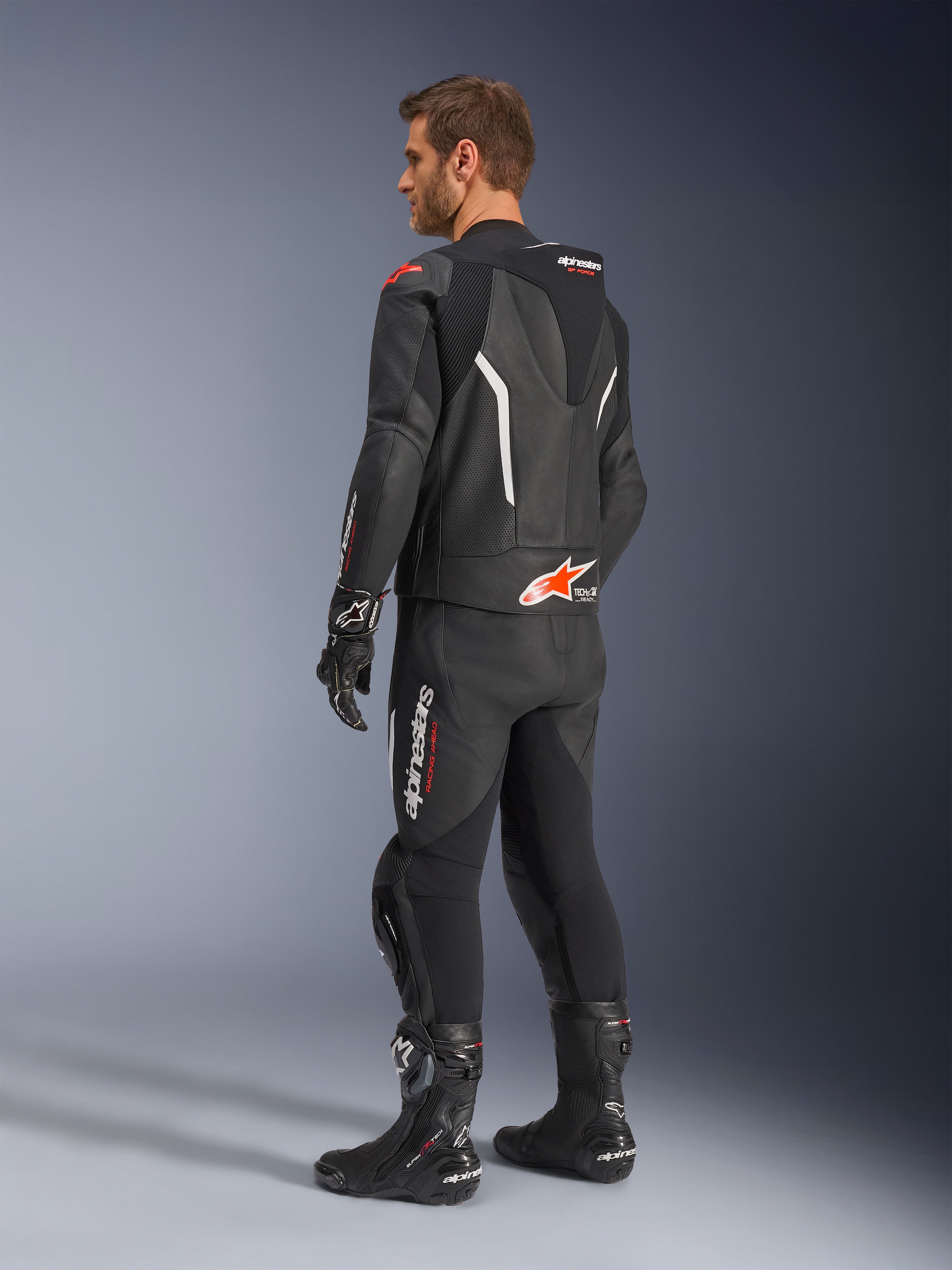 Alpinestars GP Force V2 2-teiliger Lederanzug, Schwarz mit weißen und Red Fluo Akzenten, Dreiviertel-Rückansicht am Modell, mit aerodynamischem Höcker, perforierten Ledereinsätzen und markantem Alpinestars-Branding, getragen mit schwarzen Rennstiefeln und Handschuhen.