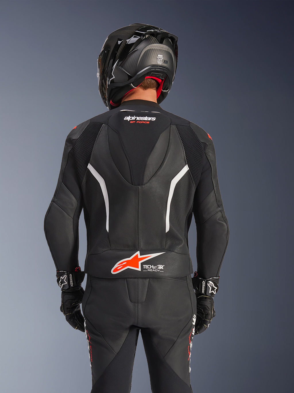Rückansicht des Alpinestars GP Force V2 2-teiligen Lederanzugs in Schwarz, mit perforiertem Leder, weißen Akzenten und rotem Alpinestars-Branding am unteren Rücken, getragen mit einem schwarzen Motorradhelm und Rennhandschuhen.