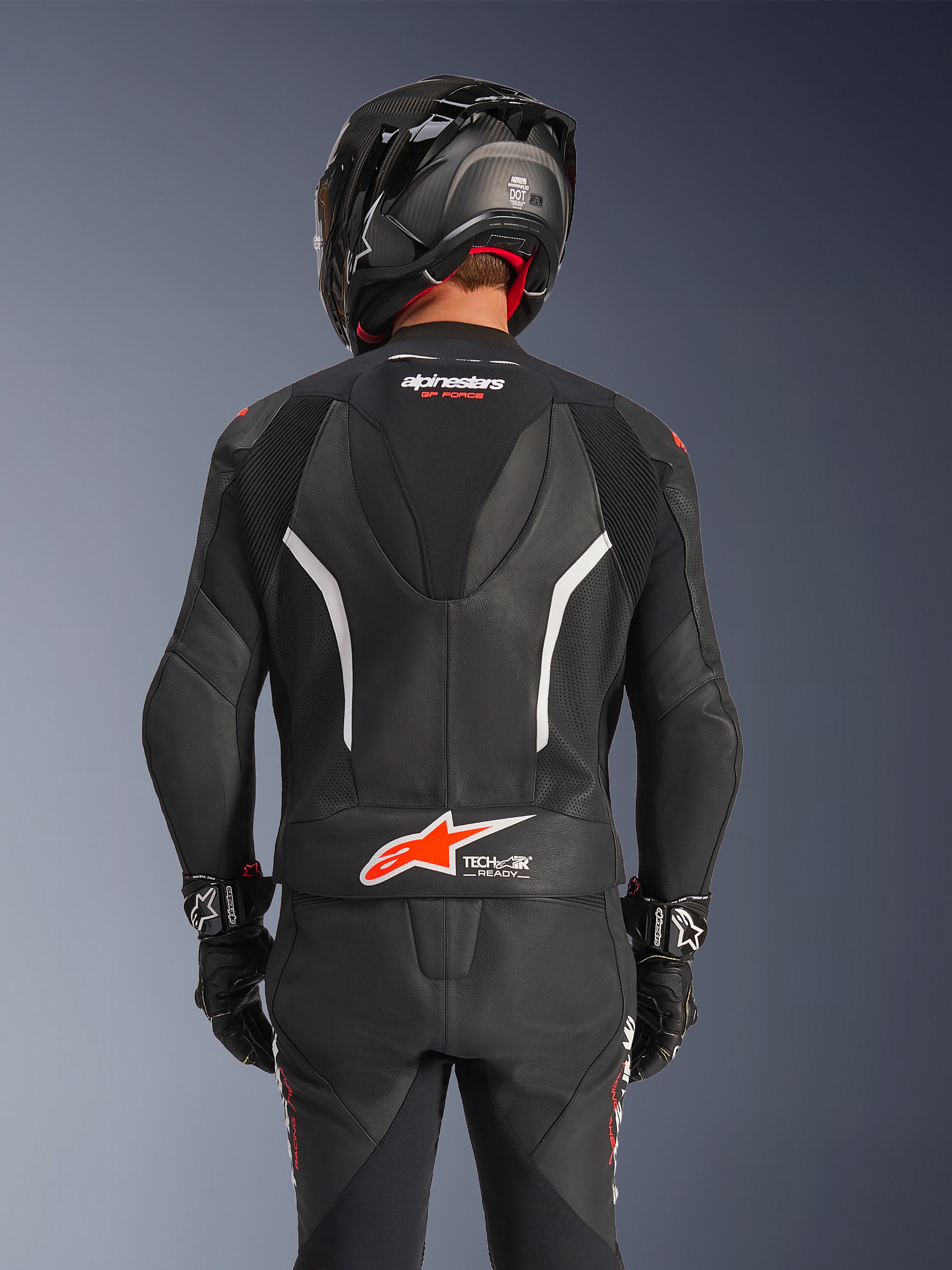 Rückansicht des Alpinestars GP Force V2 2-teiligen Lederanzugs in Schwarz, mit perforiertem Leder, weißen Akzenten und rotem Alpinestars-Branding am unteren Rücken, getragen mit einem schwarzen Motorradhelm und Rennhandschuhen.