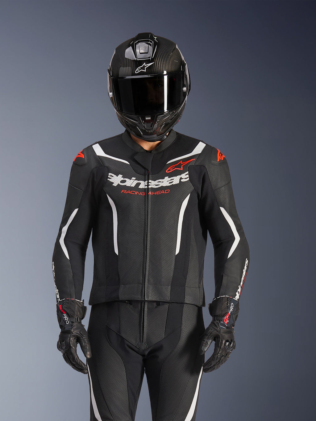 Alpinestars GP Force V2 2-teiliger Lederanzug, Schwarz, Weiß und Fluo-Rot, getragen von einem Fahrer mit einem Vollvisierhelm aus Carbon und schwarzen Rennhandschuhen, mit perforierten Ledereinsätzen und Alpinestars-Branding auf der Brust.