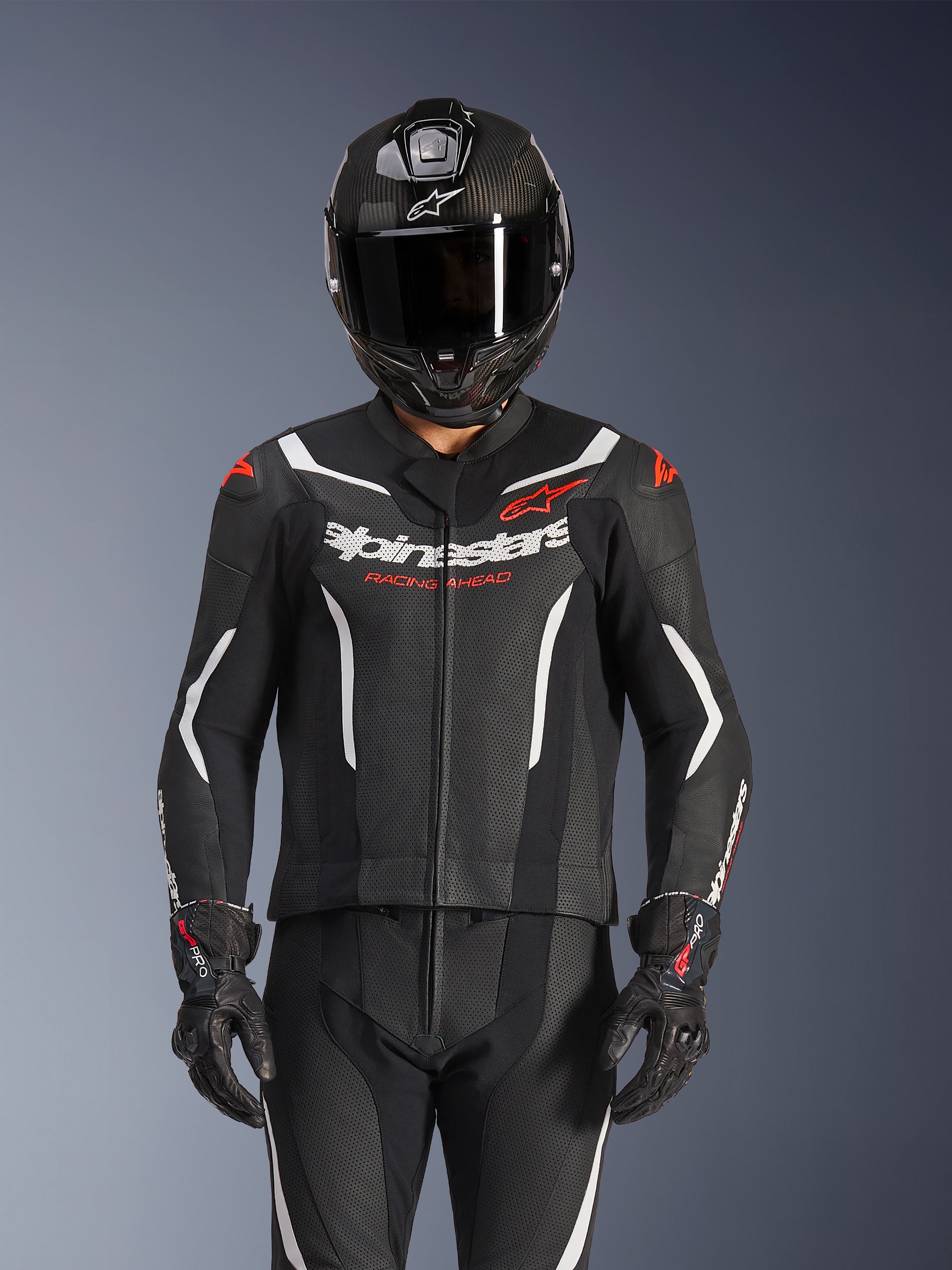 Alpinestars GP Force V2 2-teiliger Lederanzug, Schwarz, Weiß und Fluo-Rot, getragen von einem Fahrer mit einem Vollvisierhelm aus Carbon und schwarzen Rennhandschuhen, mit perforierten Ledereinsätzen und Alpinestars-Branding auf der Brust.