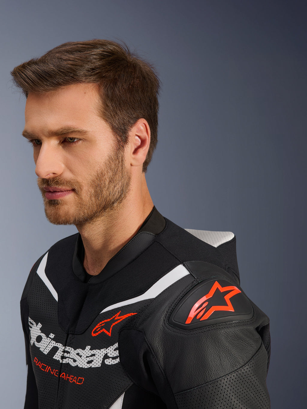Alpinestars GP Force V2 Lederkombi 2-teilig, Nahaufnahme der Jacke an einem männlichen Model, Schwarz, Weiß und Rot Fluo, mit perforiertem Leder, rotem Stern-Logo auf der Schulter, aerodynamischem Rückenhöcker und Alpinestars-Branding auf der Brust, professionelle Motorrad-Rennausrüstung