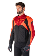 Alpinestars T-Jaws V4 Wasserdichte Jacke, Schwarz, Rot Fluo und Dunkelrot, getragen von einem männlichen Model mit schwarzer Hose und Motorradhandschuhen, aggressive sportliche Silhouette mit Alpinestars-Logo auf der Brust, Dreiviertelansicht