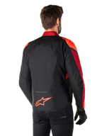 Rückansicht eines Mannes, der die Alpinestars T-Jaws V4 Wasserdichte Jacke in Schwarz Rot Fluo Dunkelrot trägt, mit leuchtend roten Akzenten an den Schultern und Ärmeln, einem fluoreszierend roten Alpinestars-Logo auf dem unteren Rücken, kombiniert mit einer schwarzen Hose und Handschuhen.