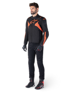 Alpinestars T-Jaws V4 Wasserdichte Jacke, Schwarz und Rot Fluo, getragen von einem männlichen Model mit schwarzer Hose, Handschuhen und Stiefeln, mit leuchtend orangefarbenen Akzenten und dem Alpinestars-Logo, sportliches Design für das Motorradfahren