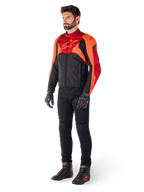 Männlicher Fahrer in der Alpinestars T-Jaws V4 Wasserdichten Jacke in Schwarz Rot Fluo Dunkelrot, kombiniert mit schwarzer Motorradhose, Handschuhen und Motorradstiefeln, sportlich geschnittenes Textildesign mit markantem Branding auf der Brust und einer aggressiven Silhouette.