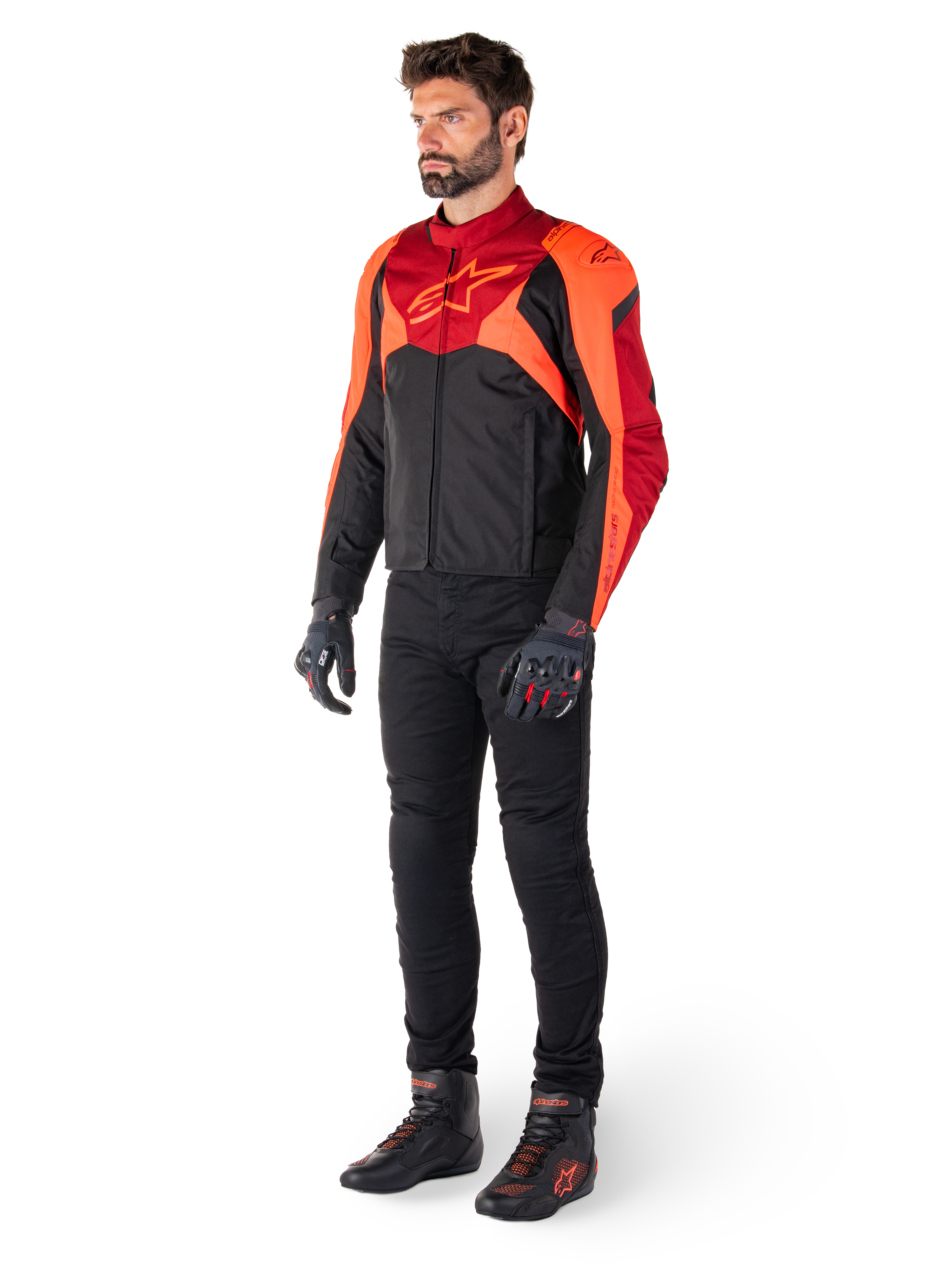 Männlicher Fahrer in der Alpinestars T-Jaws V4 Wasserdichten Jacke in Schwarz Rot Fluo Dunkelrot, kombiniert mit schwarzer Motorradhose, Handschuhen und Motorradstiefeln, sportlich geschnittenes Textildesign mit markantem Branding auf der Brust und einer aggressiven Silhouette.