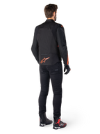 Alpinestars T-Jaws V4 Wasserdichte Jacke, Schwarz und Fluo-Rot, R&uuml;ckansicht getragen von einem m&auml;nnlichen Fahrer, mit einem leuchtend roten Fluo-Logo auf dem R&uuml;cken, kombiniert mit schwarzer Motorradhose und technischen Stiefeln, sportliche Motorradbekleidung.