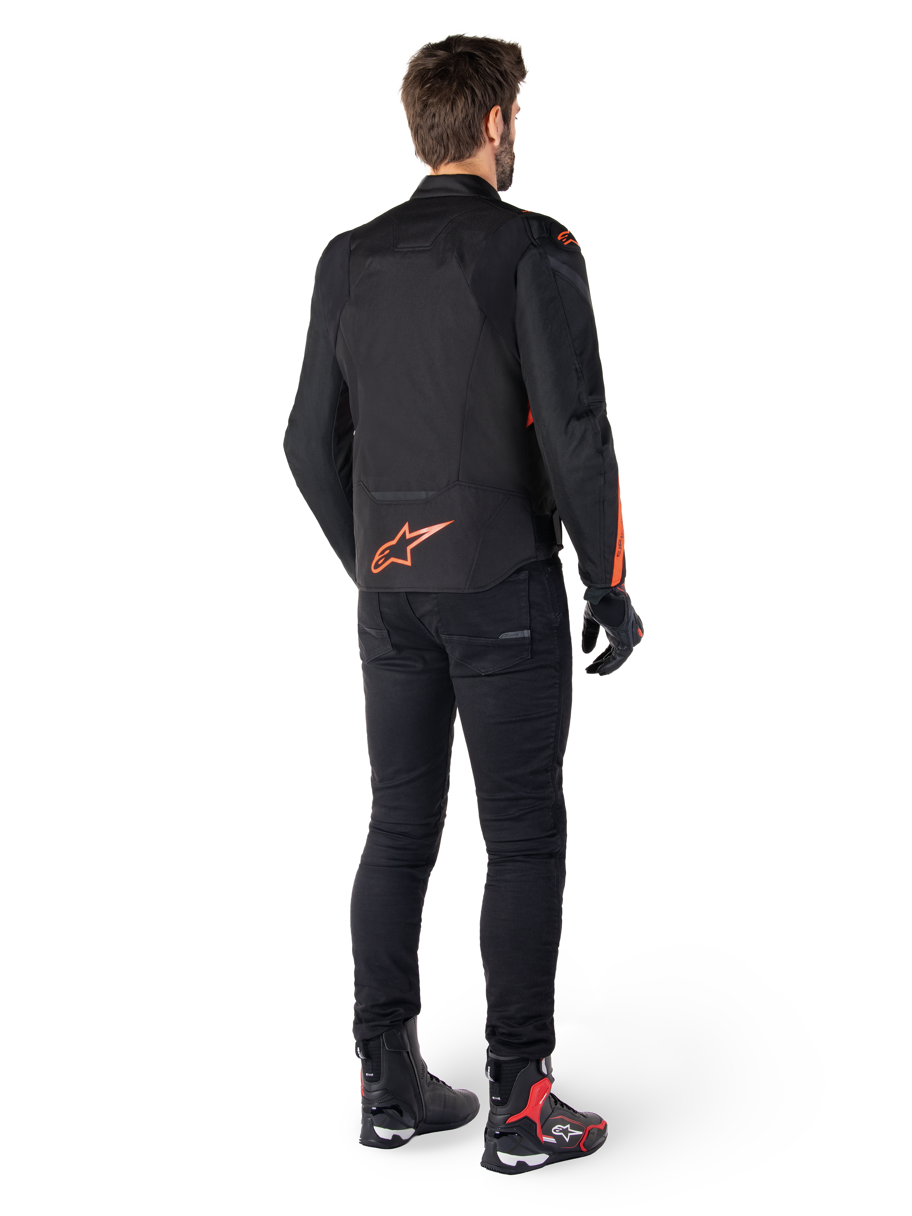 Alpinestars T-Jaws V4 Wasserdichte Jacke, Schwarz und Fluo-Rot, R&uuml;ckansicht getragen von einem m&auml;nnlichen Fahrer, mit einem leuchtend roten Fluo-Logo auf dem R&uuml;cken, kombiniert mit schwarzer Motorradhose und technischen Stiefeln, sportliche Motorradbekleidung.