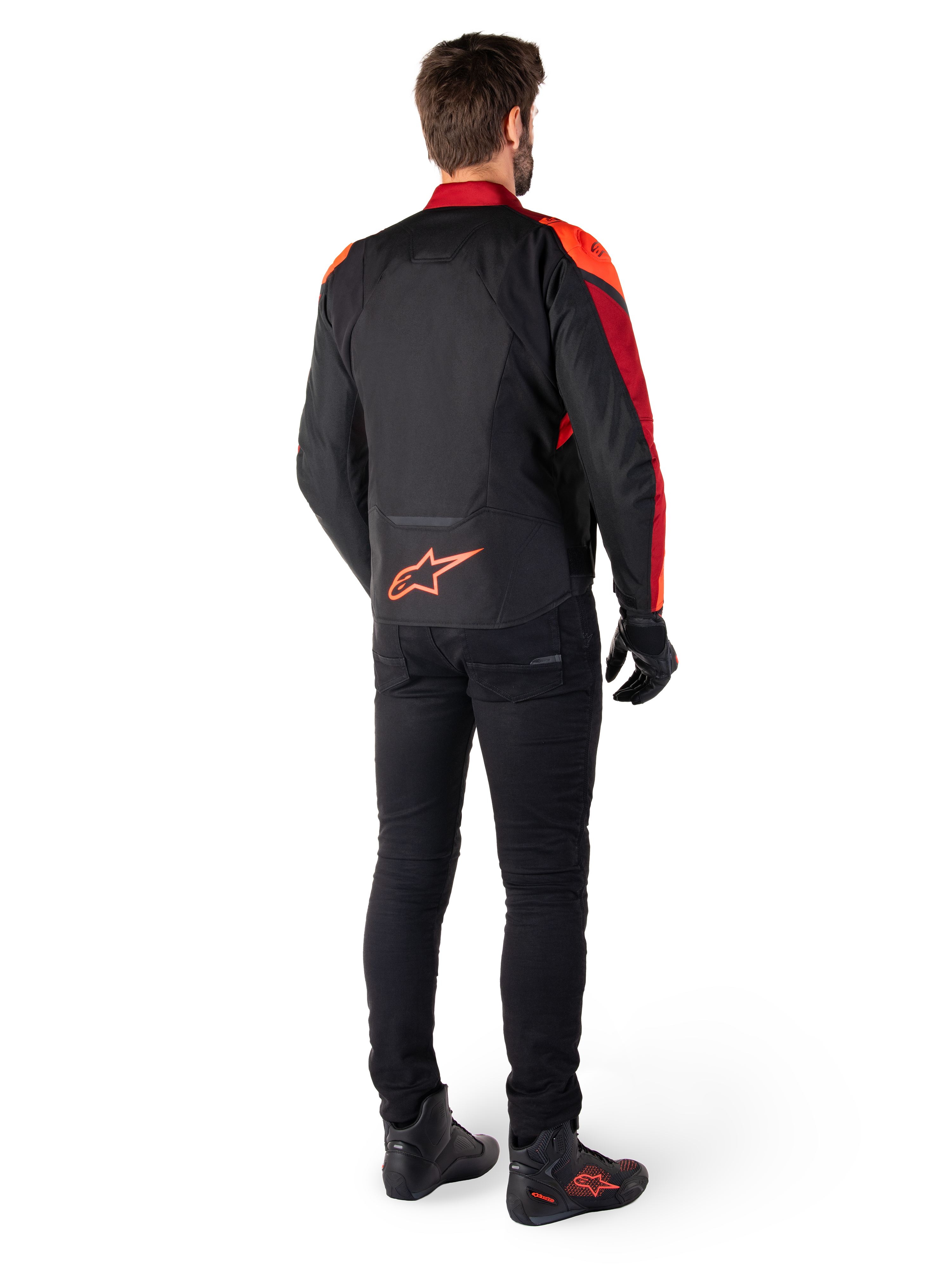 Alpinestars T-Jaws V4 Wasserdichte Jacke in Schwarz Rot Fluo Dunkelrot, Rückansicht eines Fahrers, der die Textiljacke mit schwarzer Hose und Motorradstiefeln trägt, mit einem markanten roten Alpinestars-Logo auf dem unteren Rücken.
