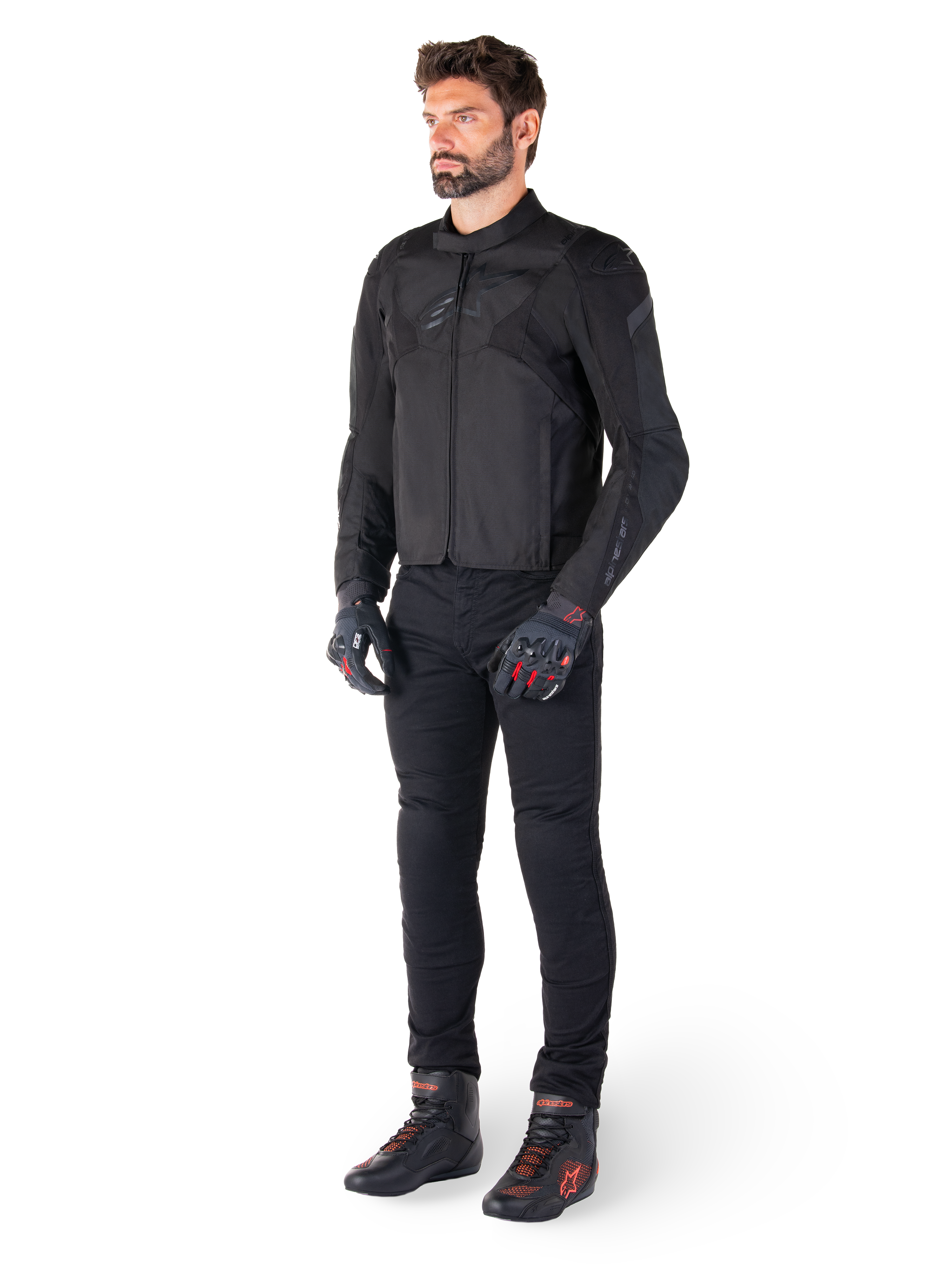 Männliches Modell in der Alpinestars T-Jaws V4 Wasserdichten Jacke in Schwarz, kombiniert mit schwarzer Motorradhose, schwarzen Handschuhen mit roten Akzenten und schwarzen Motorradschuhen, sportliche Passform, dezentes Branding, weißer Hintergrund
