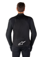Alpinestars T-Jaws V4 Waterproof Jacket, zwarte motorjas vanaf de achterkant gezien, voorzien van een wit Alpinestars-logo op de onderrug, verlengde achterzijde voor dekking, gedragen door model met zwarte broek en handschoenen, strak sportief textielontwerp