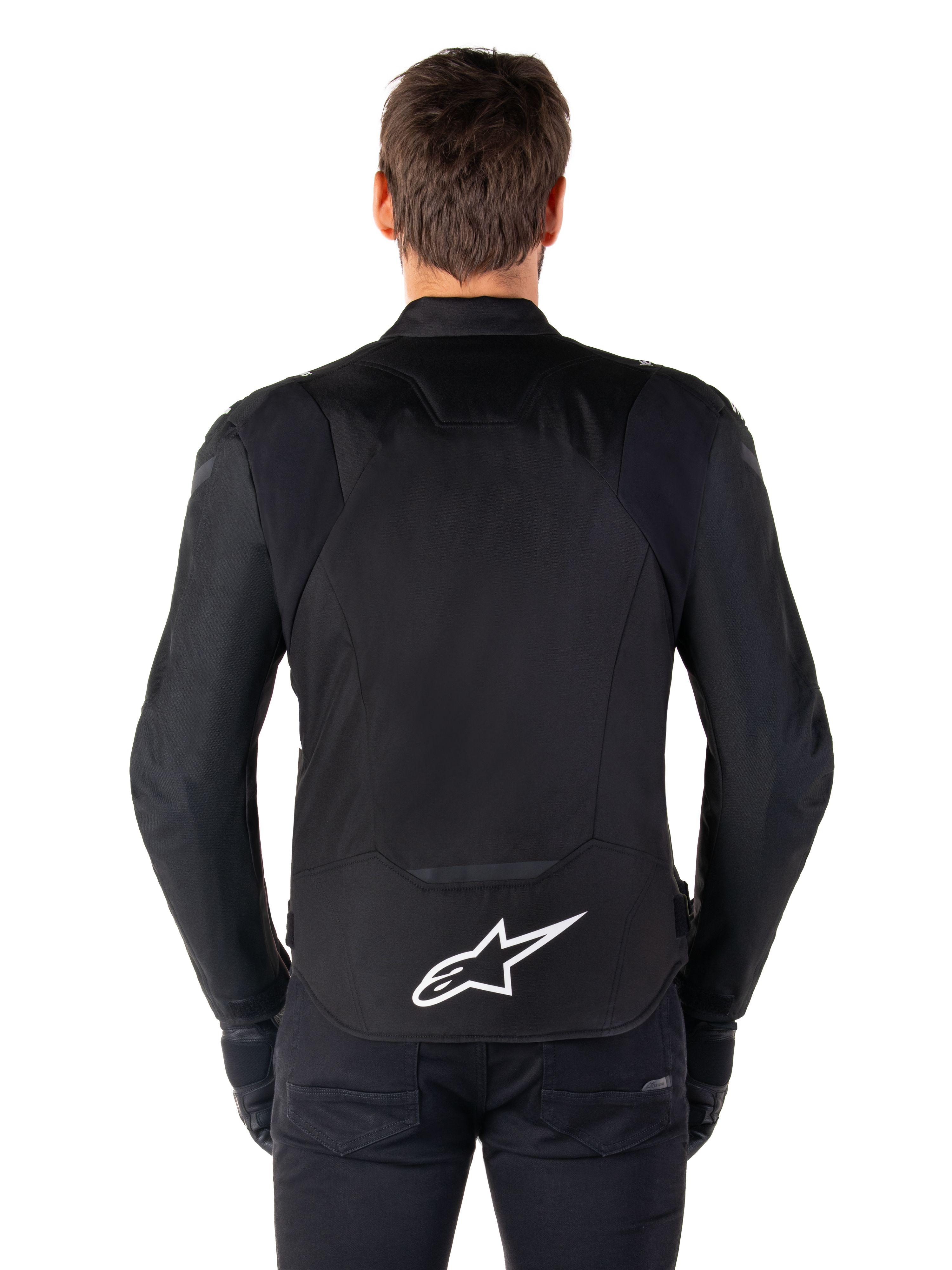 Alpinestars T-Jaws V4 Waterproof Jacket, zwarte motorjas vanaf de achterkant gezien, voorzien van een wit Alpinestars-logo op de onderrug, verlengde achterzijde voor dekking, gedragen door model met zwarte broek en handschoenen, strak sportief textielontwerp