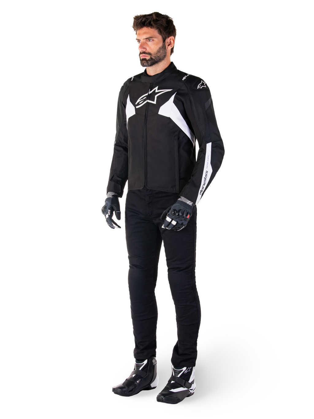 Mannelijk model draagt Alpinestars T-Jaws V4 Waterproof Jacket in zwart en wit, sportieve textieljas met een wit ster-logo en mouwaccenten, gecombineerd met een zwarte motorbroek, handschoenen en laarzen voor een complete wegrid-look