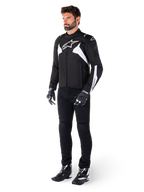 Mannelijk model draagt Alpinestars T-Jaws V4 Waterproof Jacket in zwart en wit, sportieve textieljas met een wit ster-logo en mouwaccenten, gecombineerd met een zwarte motorbroek, handschoenen en laarzen voor een complete wegrid-look