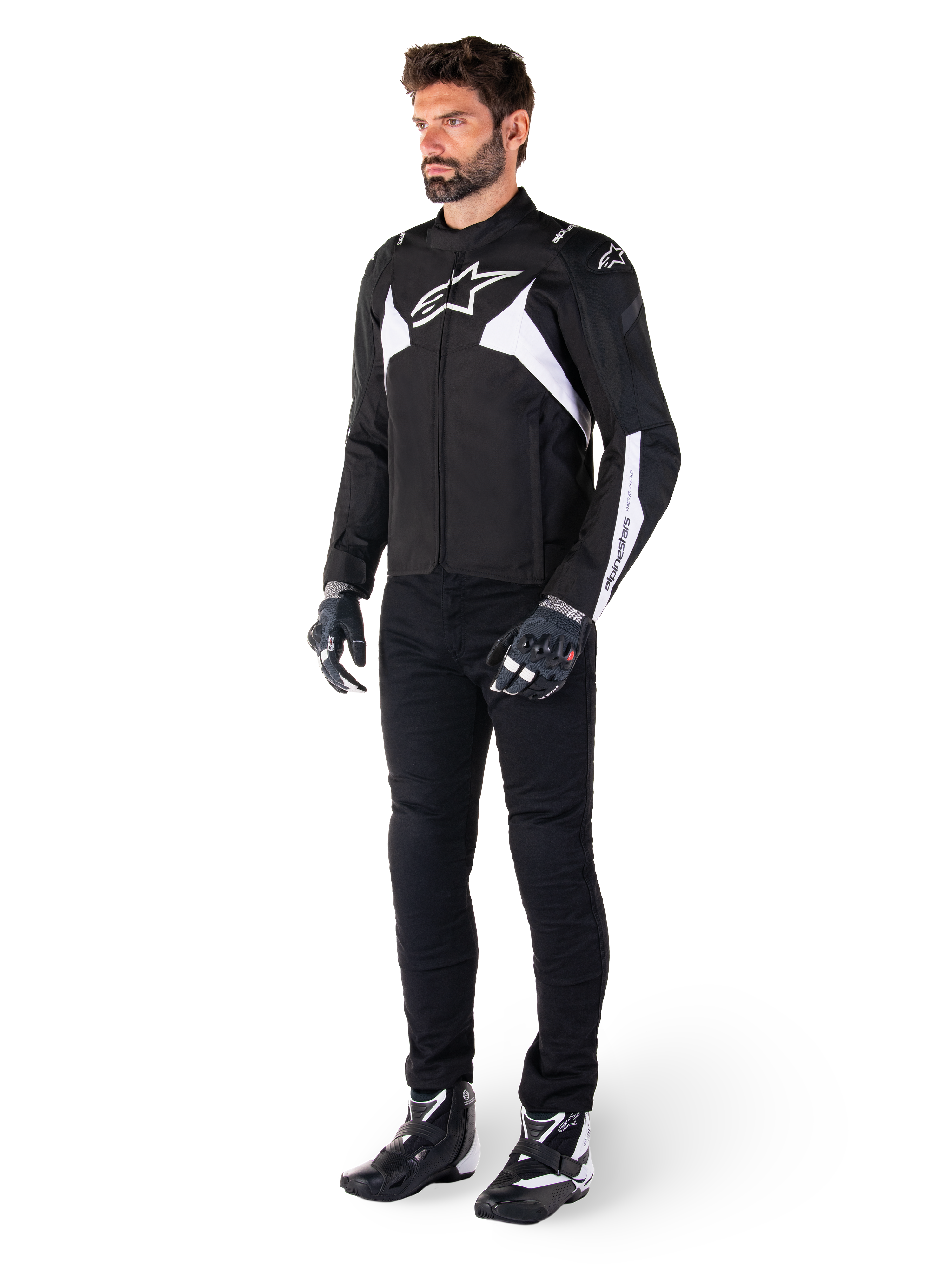 Mannelijk model draagt Alpinestars T-Jaws V4 Waterproof Jacket in zwart en wit, sportieve textieljas met een wit ster-logo en mouwaccenten, gecombineerd met een zwarte motorbroek, handschoenen en laarzen voor een complete wegrid-look