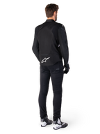 Alpinestars T-Jaws V4 Waterproof Jacket, zwart en wit, achteraanzicht met wit sterlogo op de onderrug, gedragen door model met zwarte motorbroek, zwarte handschoenen en wit-zwarte motorlaarzen