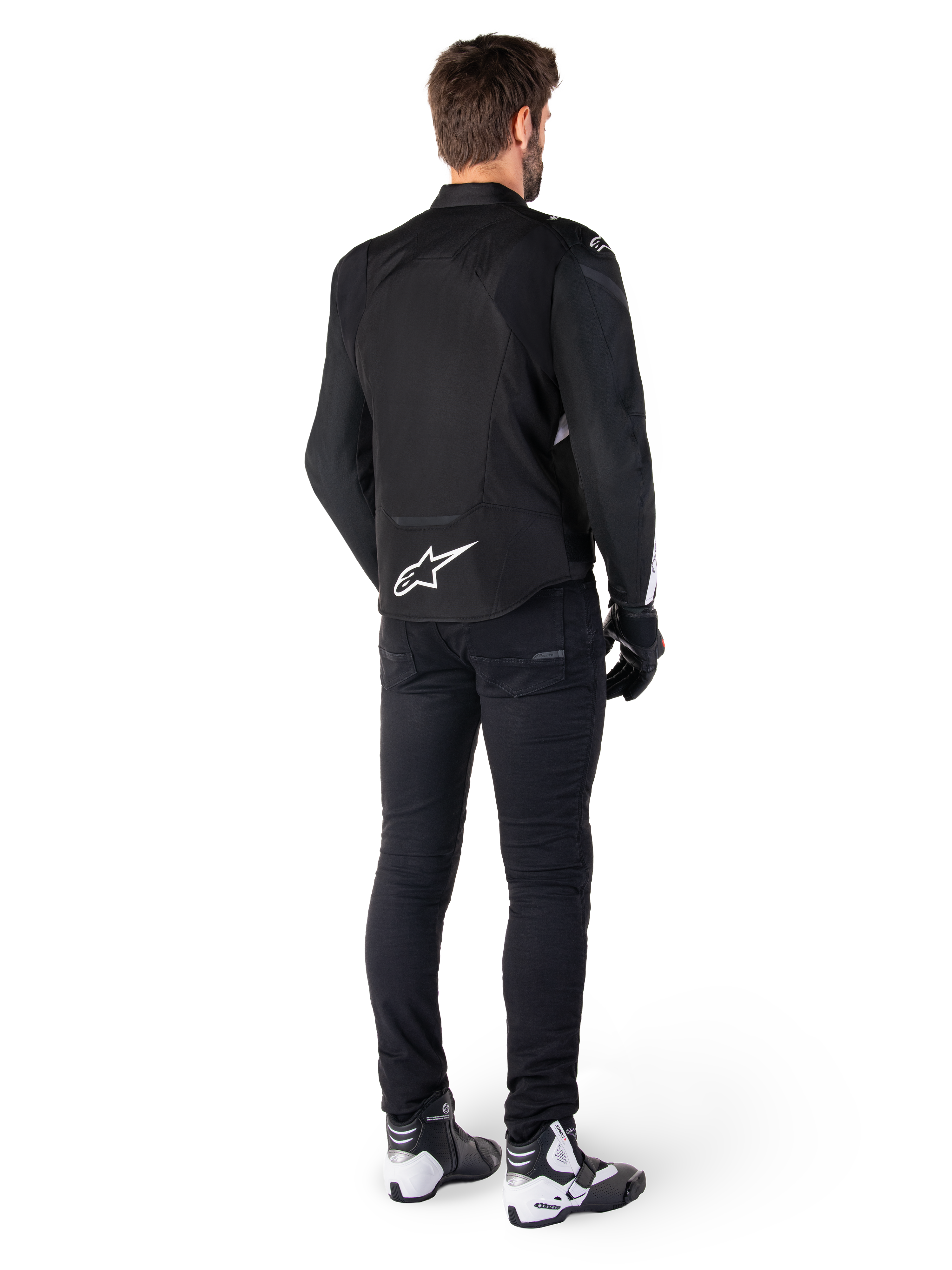 Alpinestars T-Jaws V4 Waterproof Jacket, zwart en wit, achteraanzicht met wit sterlogo op de onderrug, gedragen door model met zwarte motorbroek, zwarte handschoenen en wit-zwarte motorlaarzen