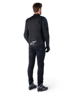 Rear view of a man wearing the Alpinestars T-Jaws V4 Waterproof Jacket in Black, Dark Blue, and Blithe Blue, featuring a prominent white Astar logo on the lower back, paired with black pants and motorcycle riding shoes.