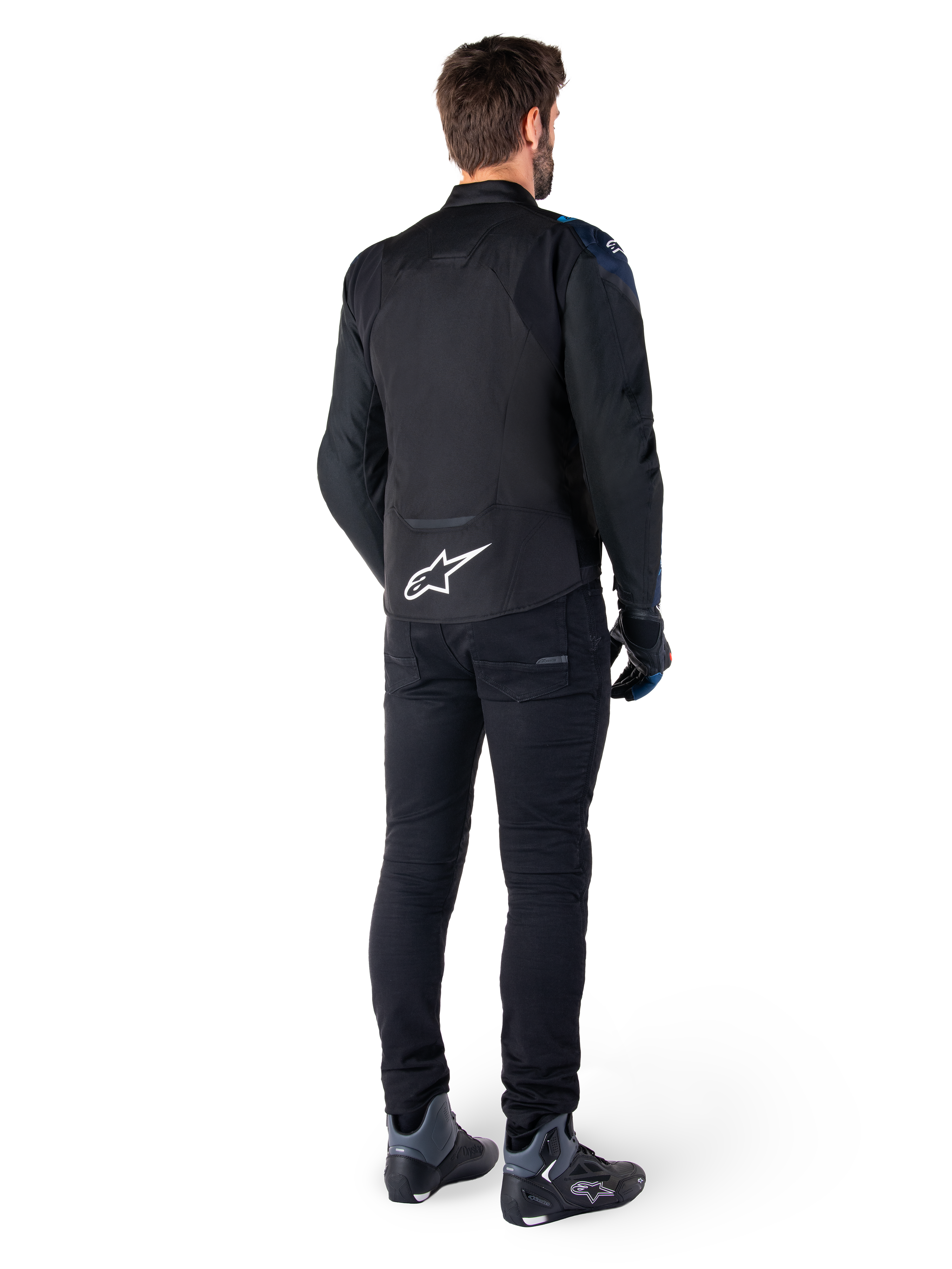 Rear view of a man wearing the Alpinestars T-Jaws V4 Waterproof Jacket in Black, Dark Blue, and Blithe Blue, featuring a prominent white Astar logo on the lower back, paired with black pants and motorcycle riding shoes.