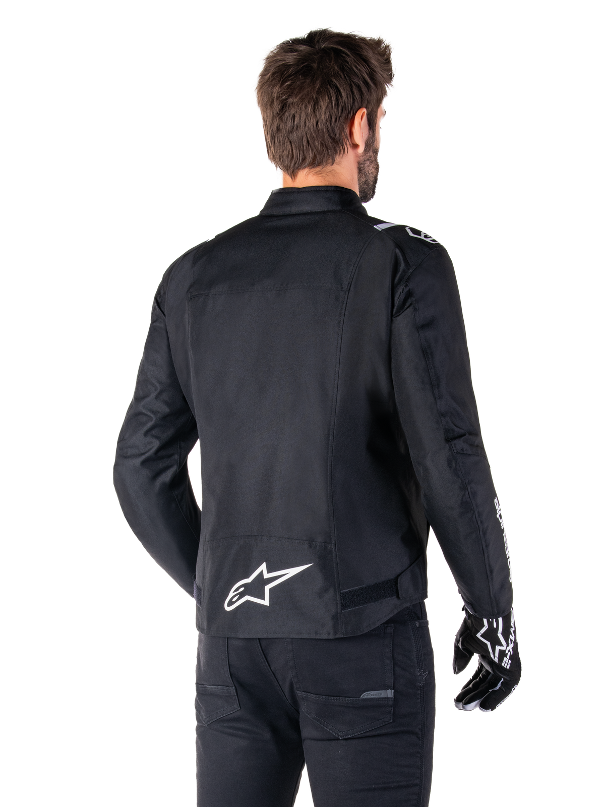 Alpinestars T-SPS V2 Wasserdichte Jacke, Schwarz und Weiß, Rückansicht eines männlichen Fahrers, der die Jacke trägt, weißes Alpinestars-Logo auf dem unteren Rücken, Textilkonstruktion im Sportschnitt, getragen mit schwarzer Motorradhose und Handschuhen