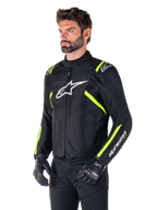 Mannequin homme portant la veste Alpinestars T-SPS V2 Waterproof, noire avec des accents blancs et jaune fluo, avec un logo Astars blanc sur la poitrine et des bandes jaune fluo sur les épaules et les manches, associée à un pantalon noir et des gants de moto, veste de moto en textile à coupe sport.