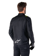 Veste imperméable Alpinestars T-SPS V2, Noir Blanc Jaune Fluo, vue arrière d'un modèle masculin portant la veste de moto en textile, avec un logo blanc en bas du dos et des accents jaune fluo sur les épaules, associée à un pantalon et des gants noirs.
