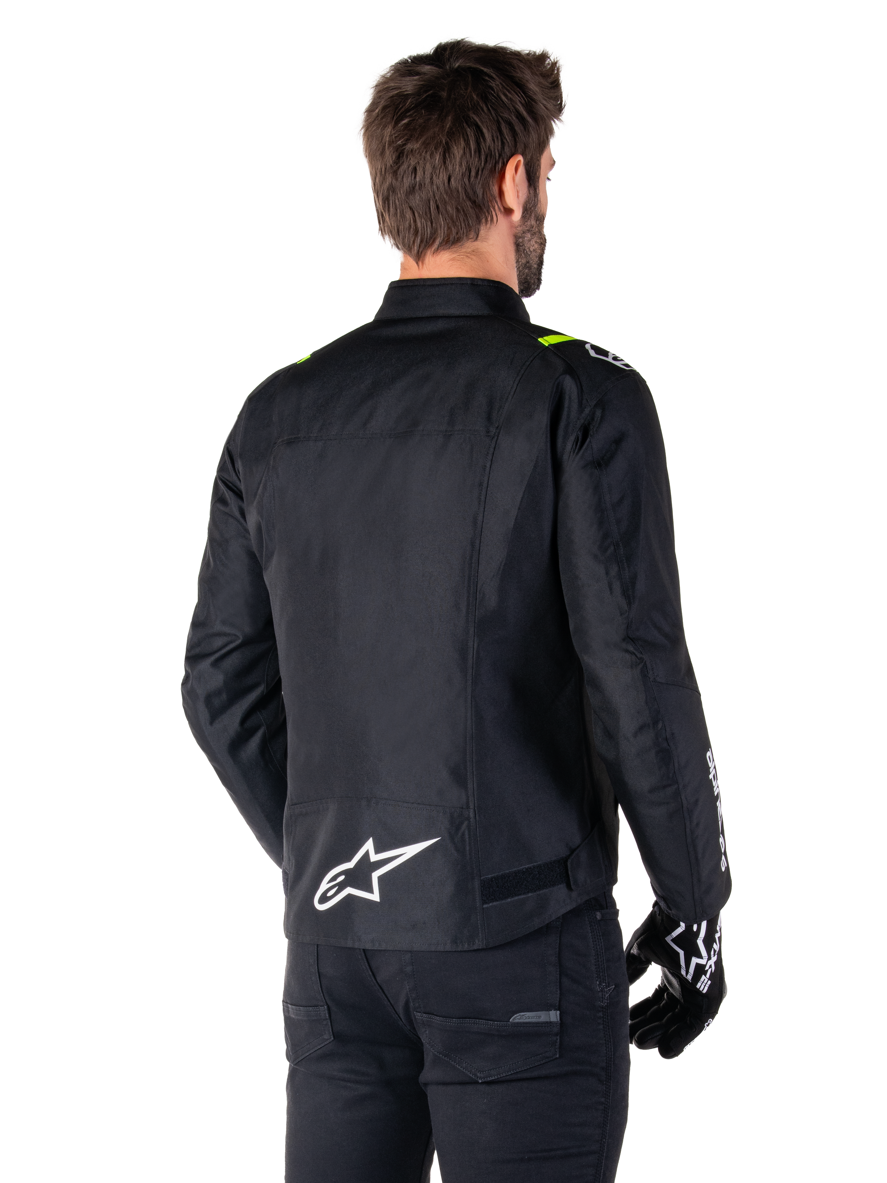 Veste imperméable Alpinestars T-SPS V2, Noir Blanc Jaune Fluo, vue arrière d'un modèle masculin portant la veste de moto en textile, avec un logo blanc en bas du dos et des accents jaune fluo sur les épaules, associée à un pantalon et des gants noirs.