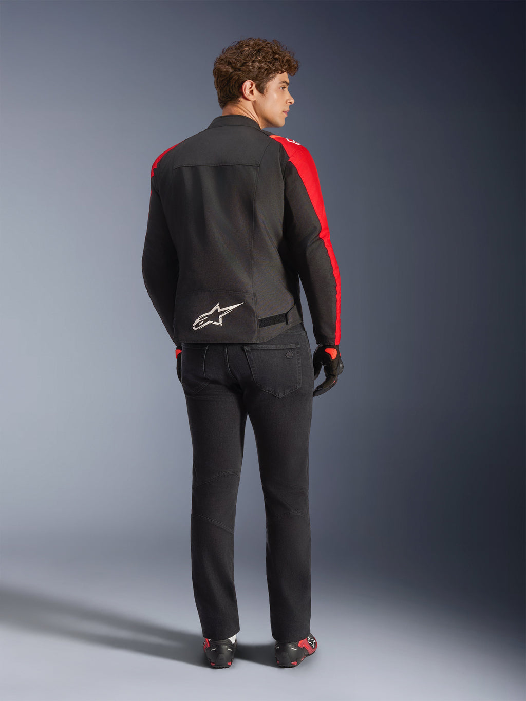 Alpinestars T-SPS V2 Wasserdichte Jacke, Schwarz Rot Rot Fluo, Rückansicht an einem männlichen Modell, weißes Alpinestars-Logo am unteren Rücken, getragen mit schwarzer Motorradhose und Handschuhen, sportliche wasserdichte Textil-Motorradjacke