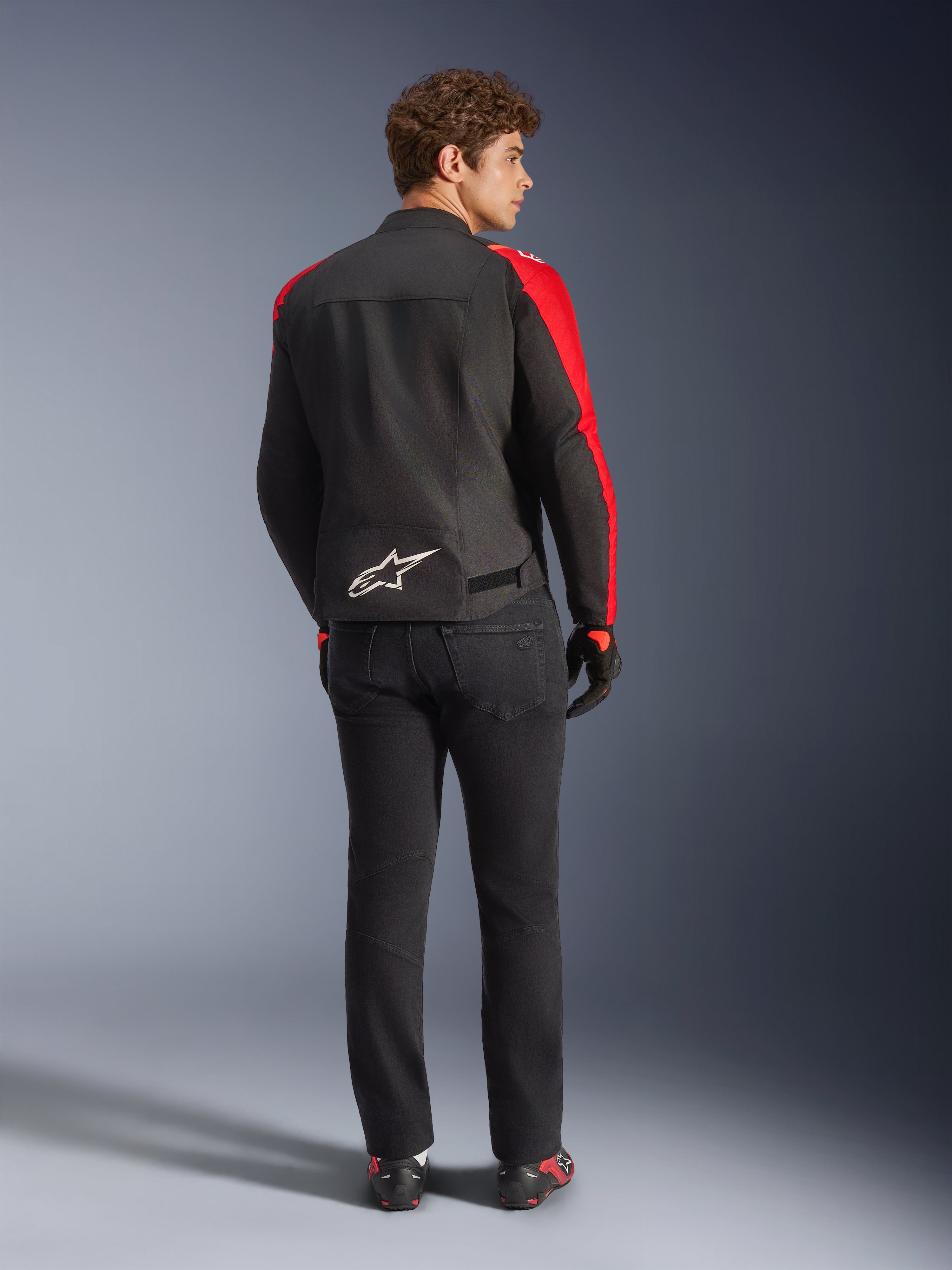 Alpinestars T-SPS V2 Wasserdichte Jacke, Schwarz Rot Rot Fluo, Rückansicht an einem männlichen Modell, weißes Alpinestars-Logo am unteren Rücken, getragen mit schwarzer Motorradhose und Handschuhen, sportliche wasserdichte Textil-Motorradjacke