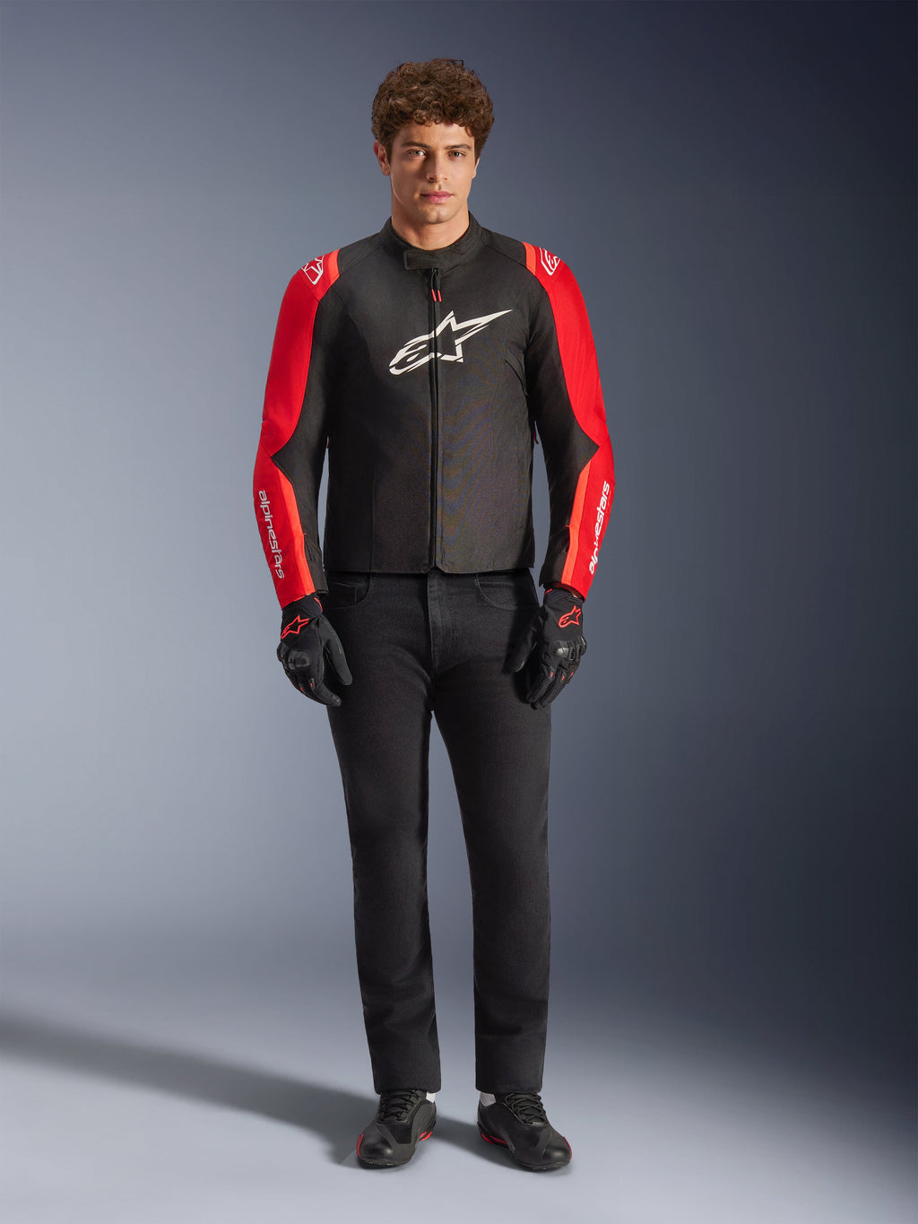 Männliches Model trägt die Alpinestars T-SPS V2 Wasserdichte Jacke, Schwarz mit roten und Red Fluo Ärmeln, weißes Logo auf der Brust, kombiniert mit schwarzer Hose, schwarzen Motorradhandschuhen und Schuhen, sportliches Motorrad-Straßenausrüstungs-Design