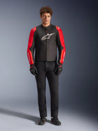 Männliches Model trägt die Alpinestars T-SPS V2 Wasserdichte Jacke, Schwarz mit roten und Red Fluo Ärmeln, weißes Logo auf der Brust, kombiniert mit schwarzer Hose, schwarzen Motorradhandschuhen und Schuhen, sportliches Motorrad-Straßenausrüstungs-Design