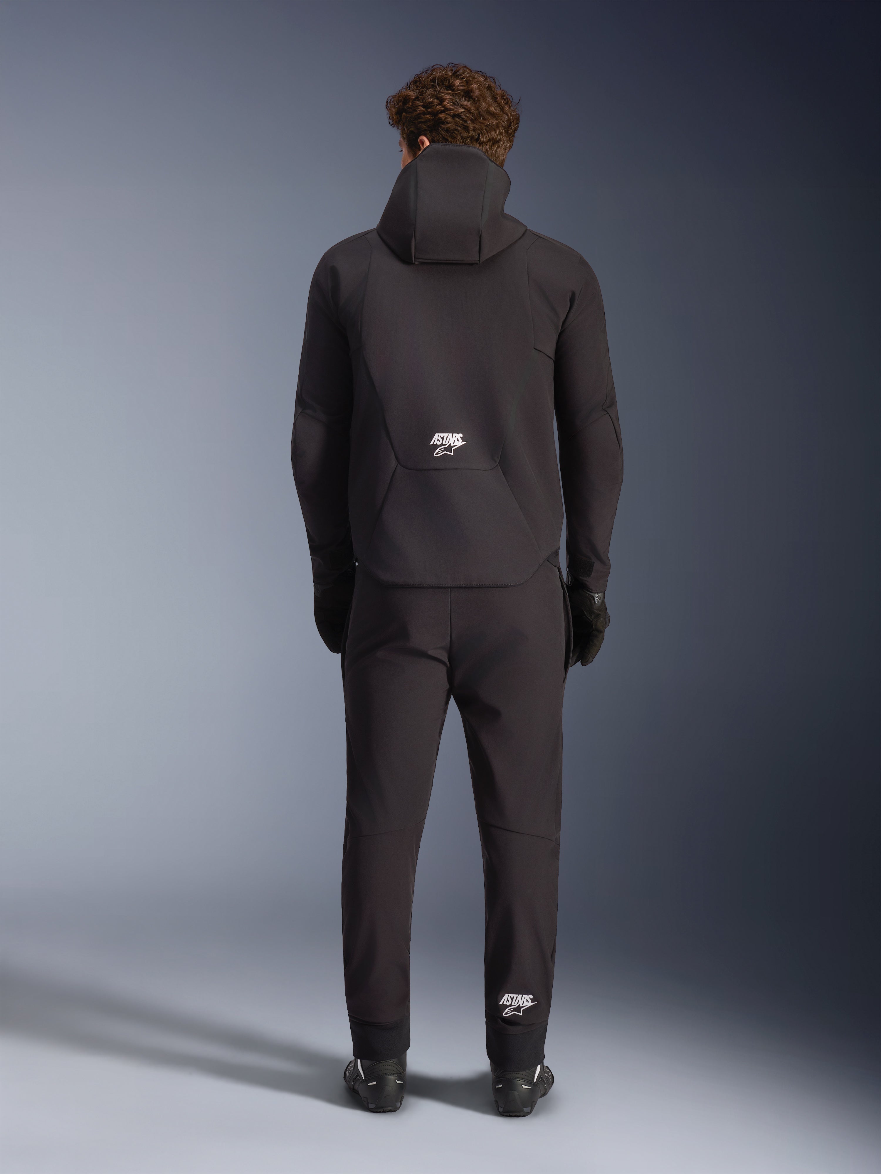 Alpinestars Aeroshell Waterproof Jacke, Schwarz, Rückansicht an einem Model in passender schwarzer Motorradhose und Handschuhen, Sport-Softshell-Design mit Kapuze und weißen ASTARS-Logos, elegante und moderne Motorradbekleidung