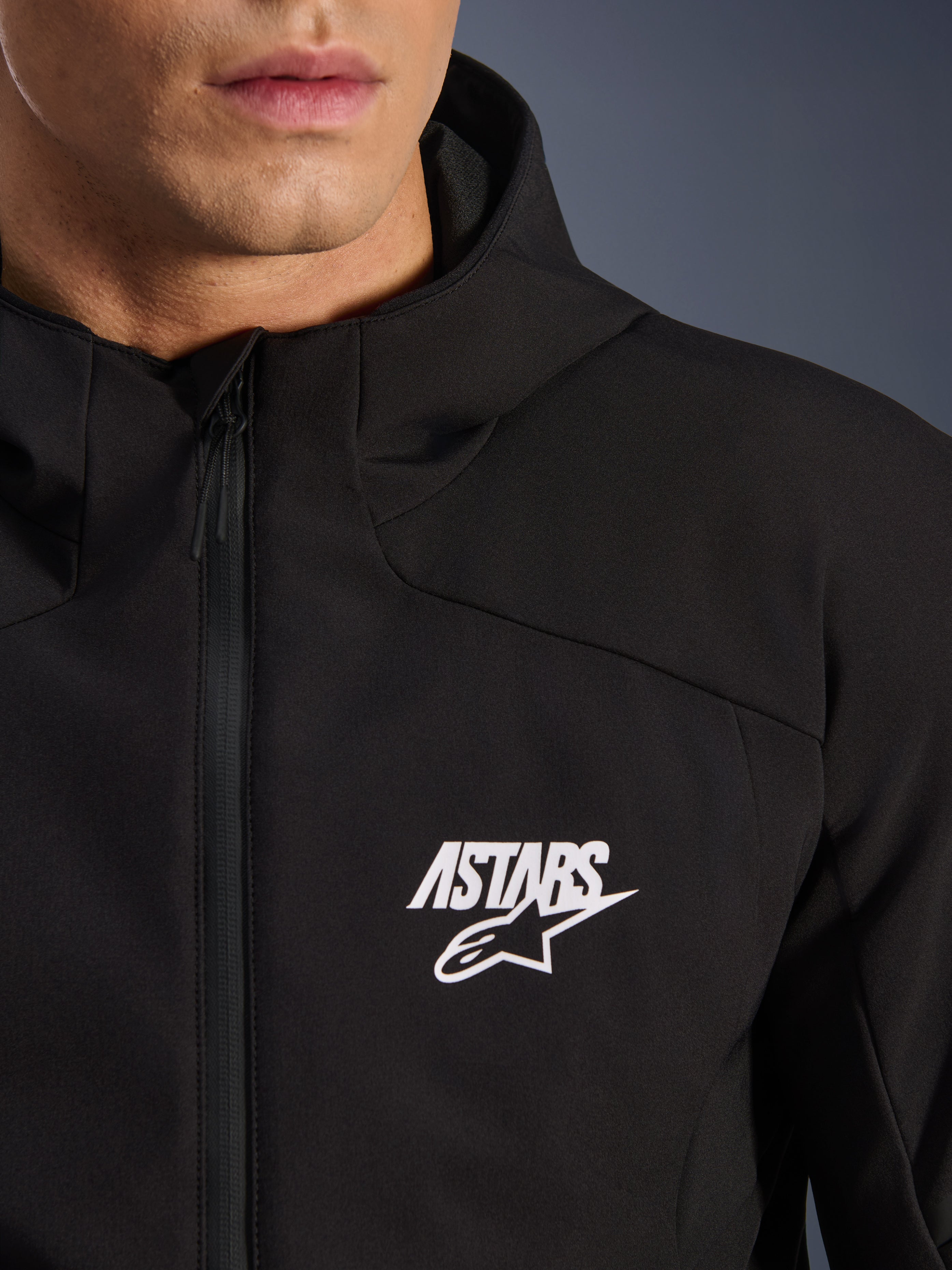 Alpinestars Aeroshell Waterproof Jacke, schwarze Motorrad-Softshelljacke, Nahaufnahme des Brustbereichs mit weißem ASTARS Stern-Logo und wasserdichtem Frontreißverschluss, integrierte Kapuze, modernes sportliches Design