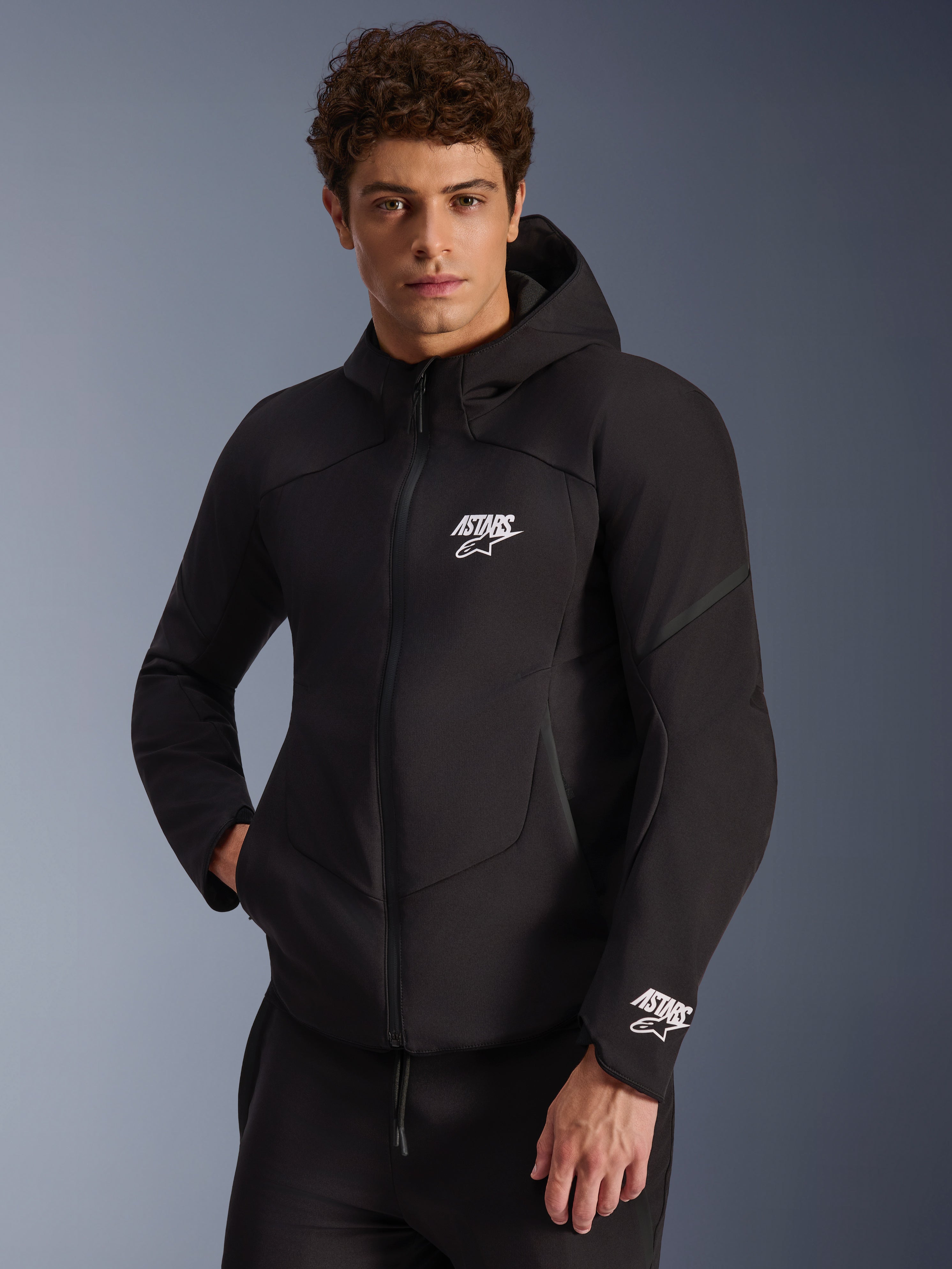 Alpinestars Aeroshell WP Jacke, schwarzer Softshell-Motorrad-Hoodie, weißes ASTARS-Logo auf Brust und Ärmelbündchen, schlankes Design mit integrierter Kapuze und wasserdichtem Reißverschluss, moderne sportlich inspirierte Ausrüstung für urbane Fahrten