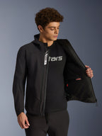 Alpinestars Aeroshell Waterproof Jacke, schwarze Softshell-Motorradjacke, geöffnet mit sichtbarer Innentasche und Innenfutter, getragen von einem männlichen Model mit schwarzem Logo-T-Shirt, Kapuzendesign für lässigen urbanen Fahrstil und Street-Style.