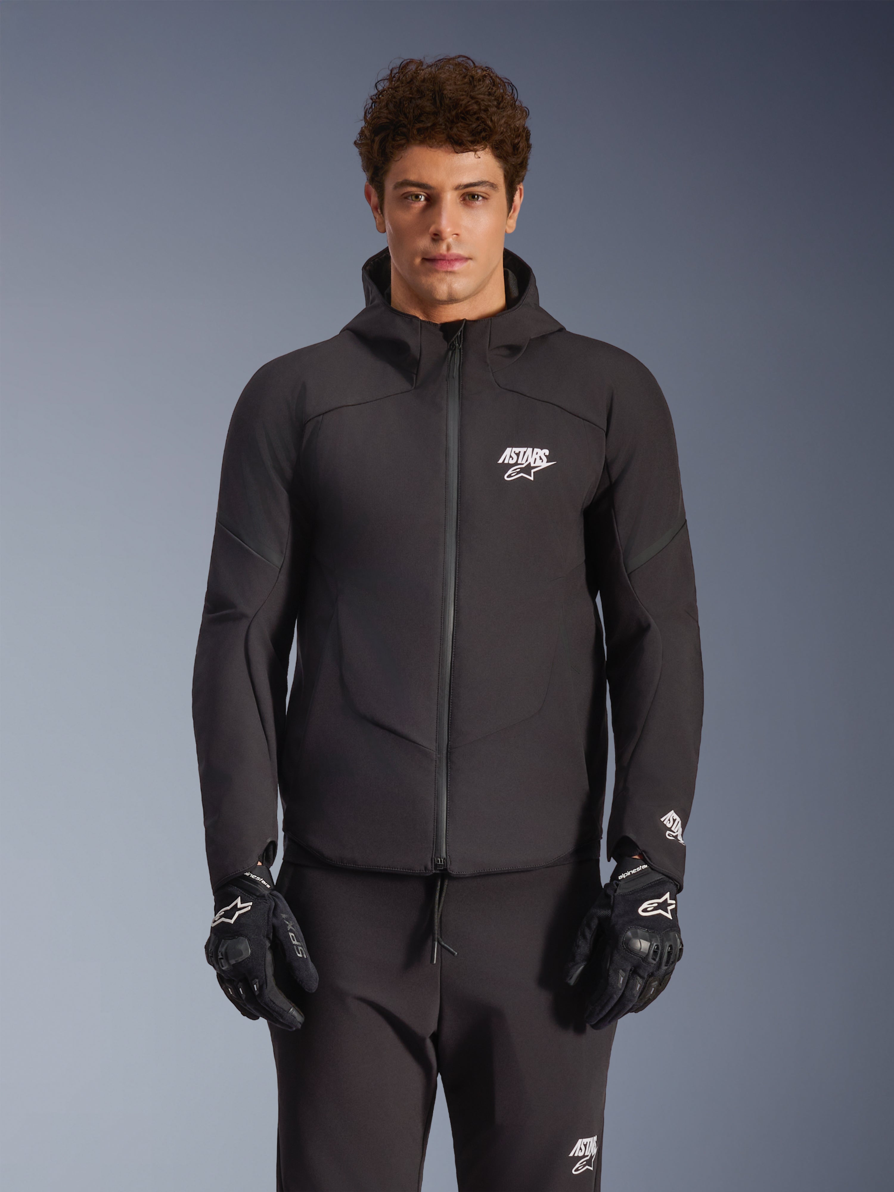Alpinestars Aeroshell Waterdichte Jas, zwarte textiel softshell urban motorjas voor heren met een geïntegreerde capuchon, gedragen met zwarte motorhandschoenen.