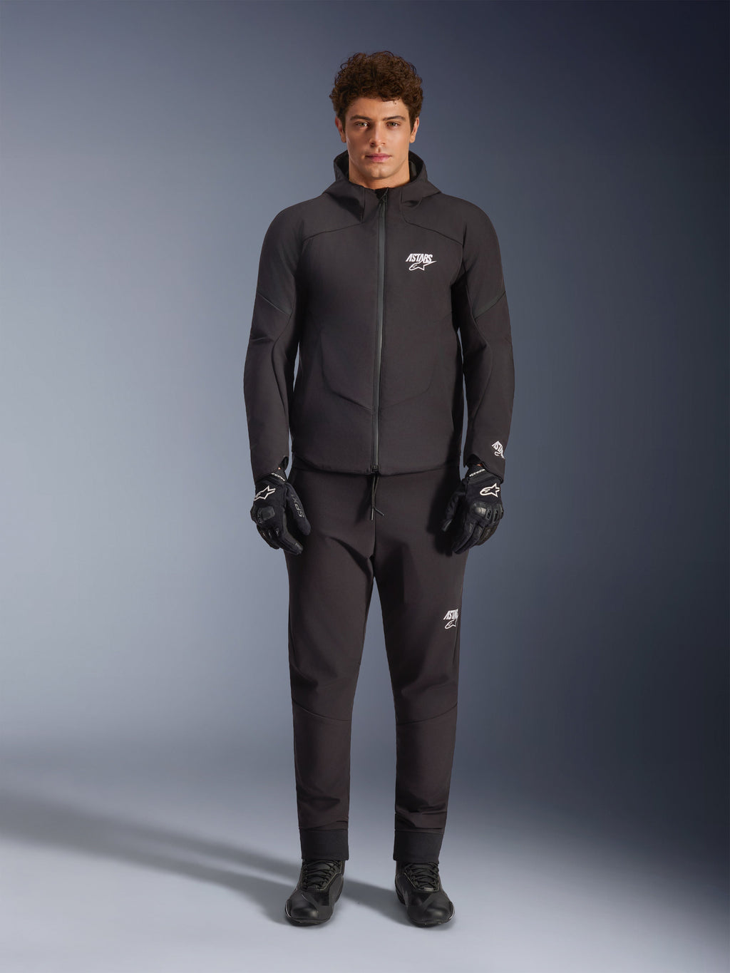 Männliches Model in schwarzer Alpinestars Aeroshell Waterproof Jacke, kombiniert mit passender technischer Hose und Motorradhandschuhen, Softshell-Design mit Kapuze und weißen Logo-Akzenten, modernes, vom Motorsport inspiriertes Outfit für die Straße