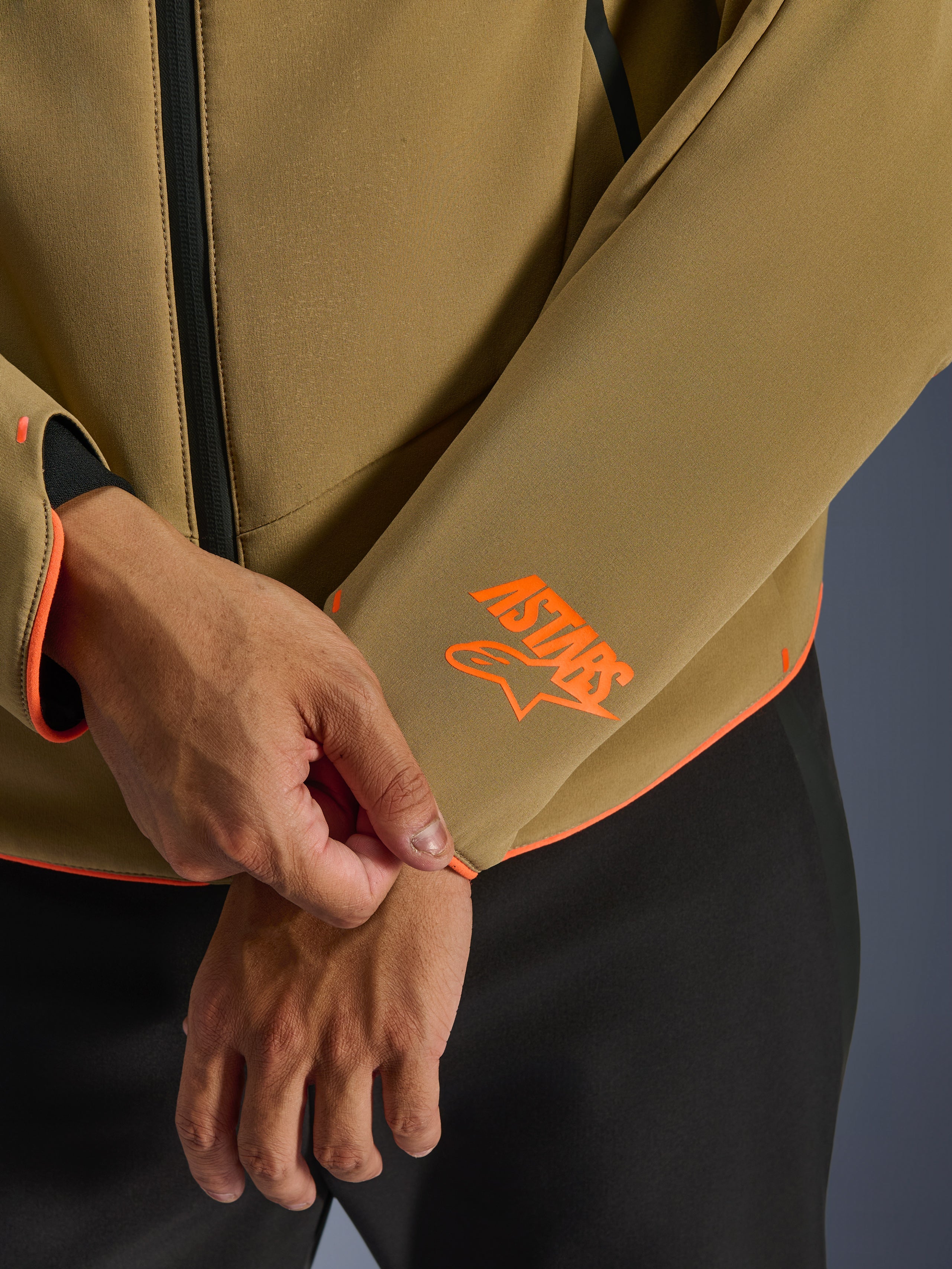 Alpinestars Aeroshell Waterproof Jacket, Dark Khaki with Orange Fluo accents, close-up of the sleeve cuff and lower hem featuring the bright orange Astars logo, neon orange piping, and waterproof front zipper detail, sport softshell construction.