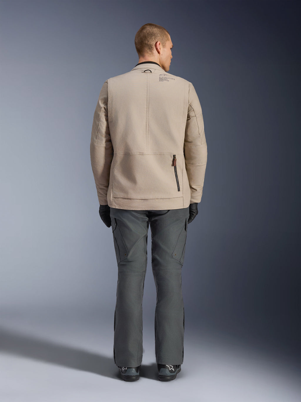 Veste Alpinestars Borrego Drystar®, vue arrière d'un mannequin homme portant la veste de moto textile Warm Gray, associée à un pantalon technique gris foncé, des gants noirs et des bottes, présentant le design robuste en toile cirée et des panneaux d'extension à l'arrière.
