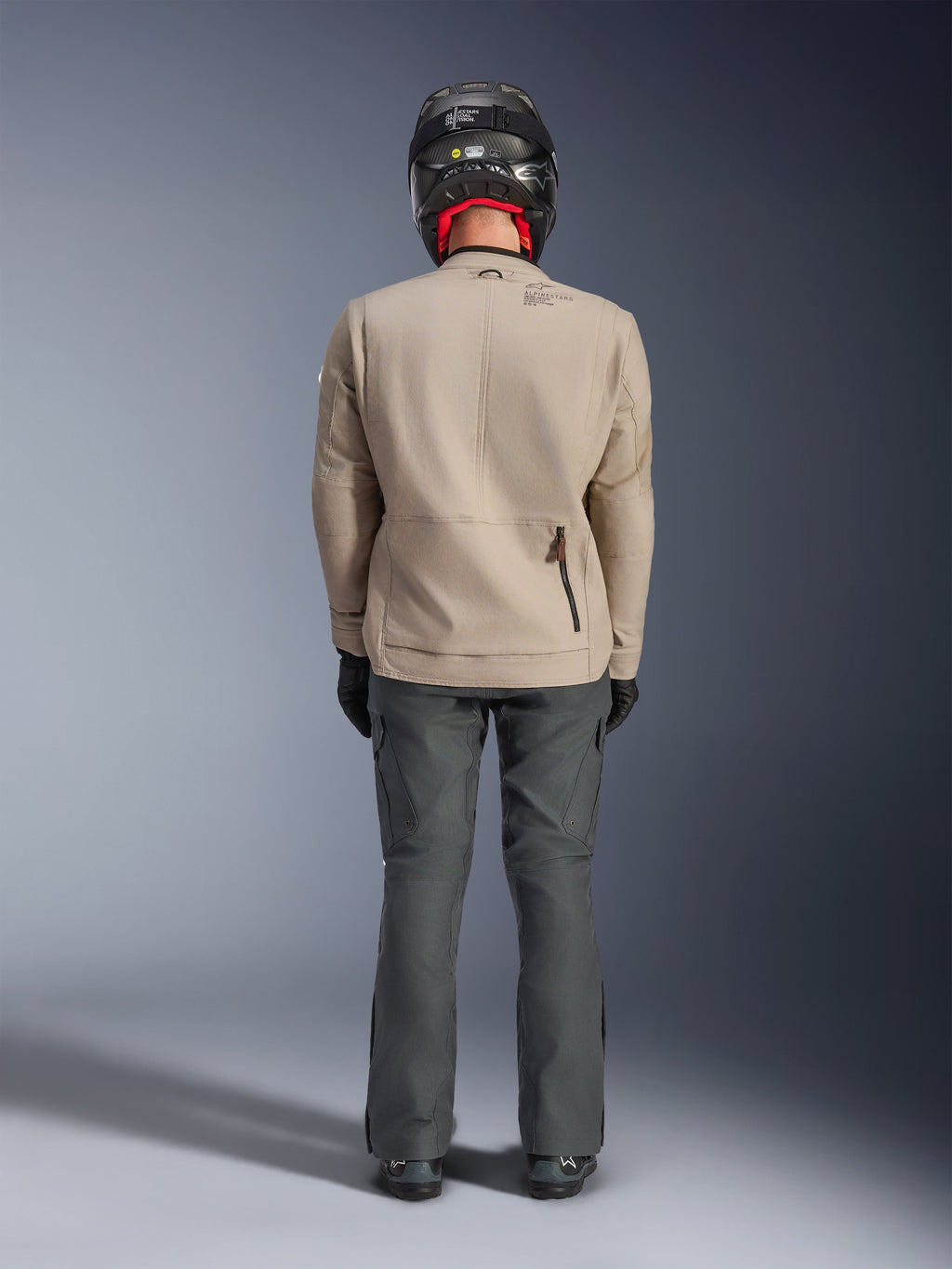 Veste Alpinestars Borrego Drystar®, veste de moto d'aventure gris chaud, vue arrière montrant un aspect toile cirée avec panneaux d'expansion aux épaules et poche zippée au bas du dos, portée avec un pantalon de moto gris foncé et un casque noir.