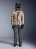 Veste Alpinestars Borrego Drystar® en Warm Gray, veste de moto d'aventure avec quatre poches cargo à l'avant, associée à un pantalon de moto gris foncé, un casque tout-terrain noir et des gants noirs, présentant un aspect toile cirée robuste