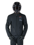 Alpinestars AMT-8 Stretch Drystar® XF Jacke, schwarze Motorradjacke, mit orangefarbenem Alpinestars-Logo und grauem AMT-Branding, hoher Kragen mit Frontreißverschluss und Belüftungsöffnungen, getragen mit schwarzem Helm und Handschuhen, schützende Adventure-Tourenbekleidung